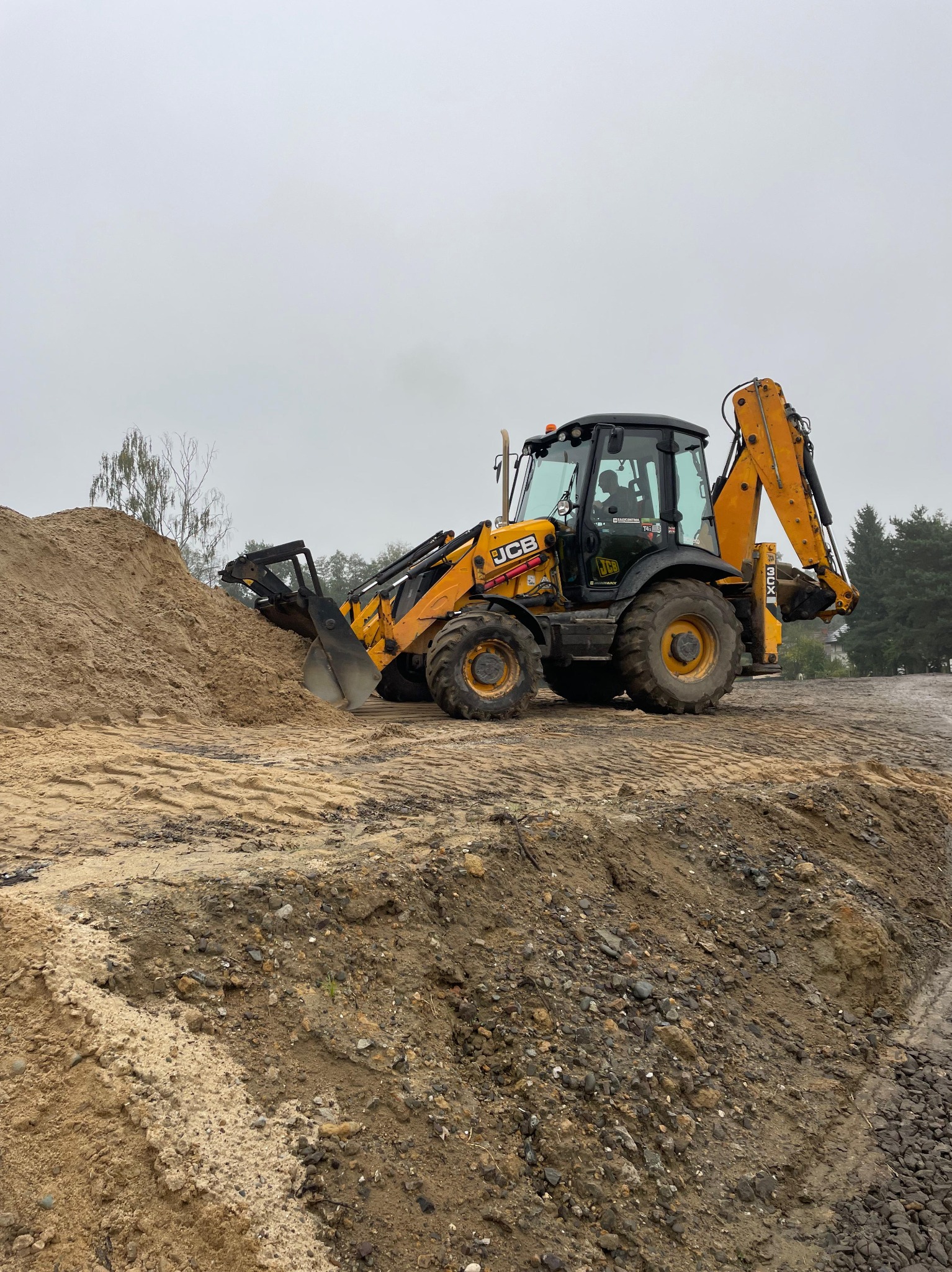 Żółto-czarna koparko-ładowarka JCB na placu budowy, obok pryzma piasku. Widoczne ślady opon na ziemi. Pochmurne niebo w tle.