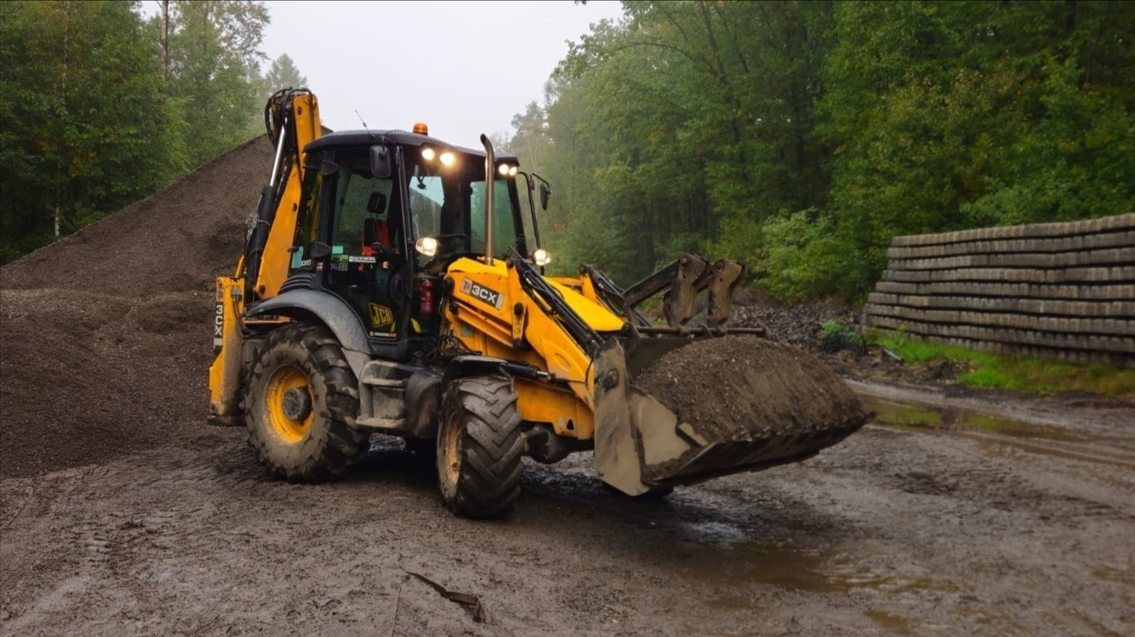 Żółta koparko-ładowarka JCB 3CX z pełną łyżką ziemi na błotnistym terenie, w tle hałda żwiru i ściana z betonowych prefabrykatów, otoczona zielenią.
