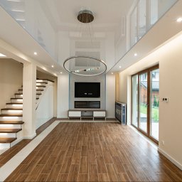 SENDO - Salon z drewnopodobną podłogą, schodami z podświetleniem i nowoczesnym oświetleniem sufitowym. Widoczne duże okno balkonowe i telewizor na ścianie.