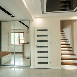 SENDO - Jasne wnętrze domu z drewnianymi schodami podświetlonymi LED, białymi drzwiami z przeszkleniami i lustrzaną szafą wnękową. Widoczna kuchnia w tle.