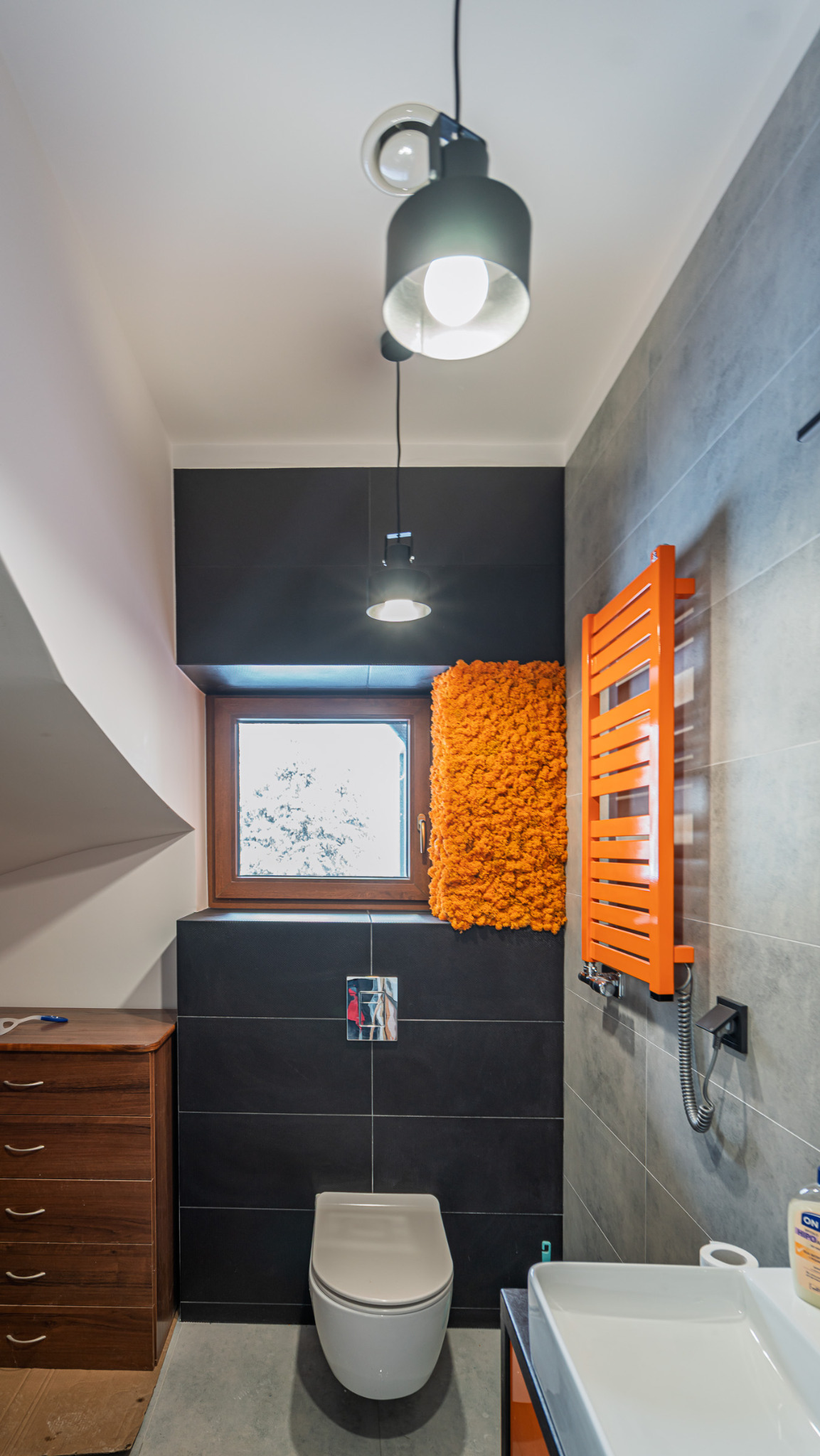 Nowoczesna łazienka na poddaszu z czarnymi i szarymi płytkami, pomarańczowym grzejnikiem i mchem dekoracyjnym przy oknie. Minimalistyczne oświetlenie.
