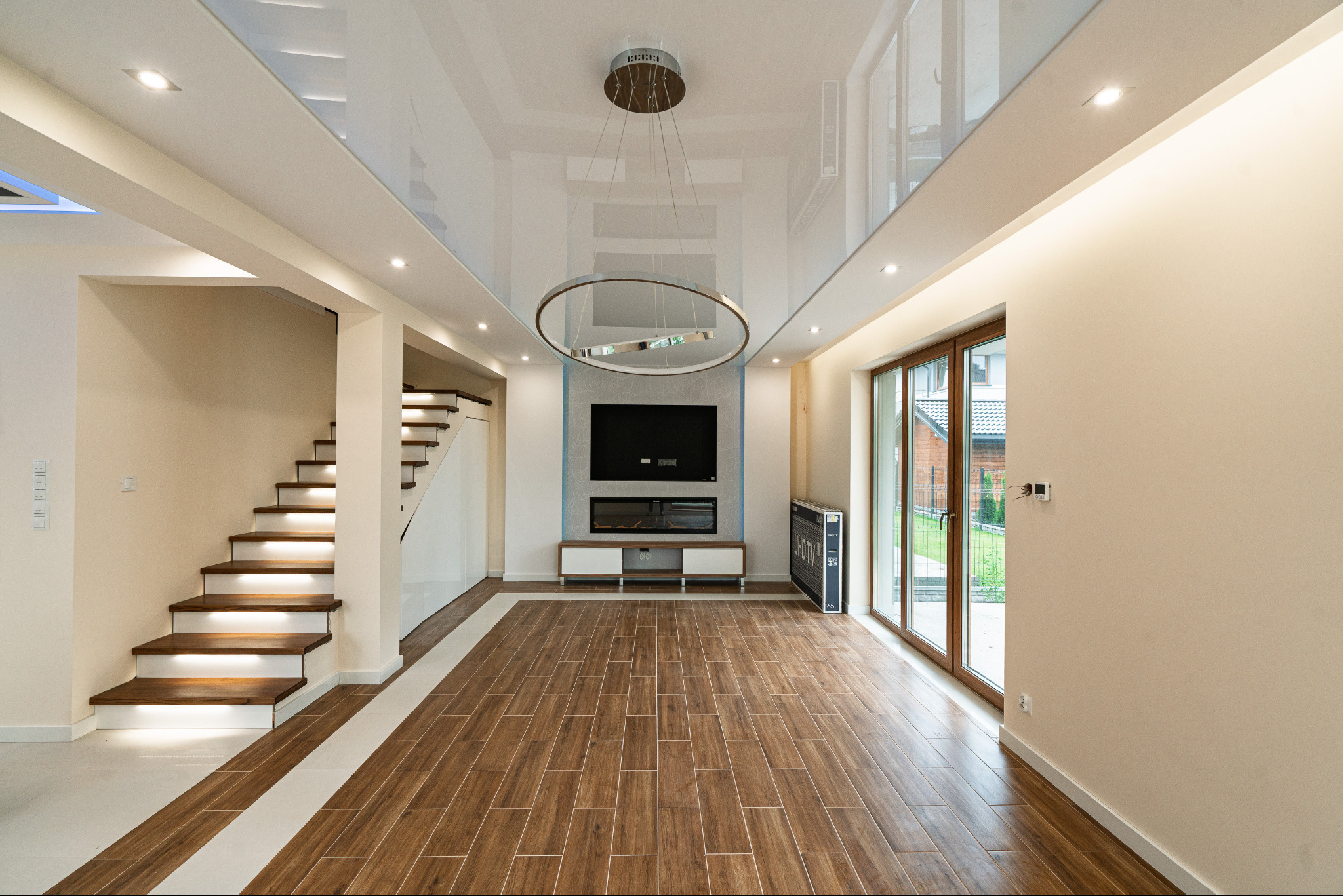 Salon z drewnopodobną podłogą, schodami z podświetleniem i nowoczesnym oświetleniem sufitowym. Widoczne duże okno balkonowe i telewizor na ścianie.