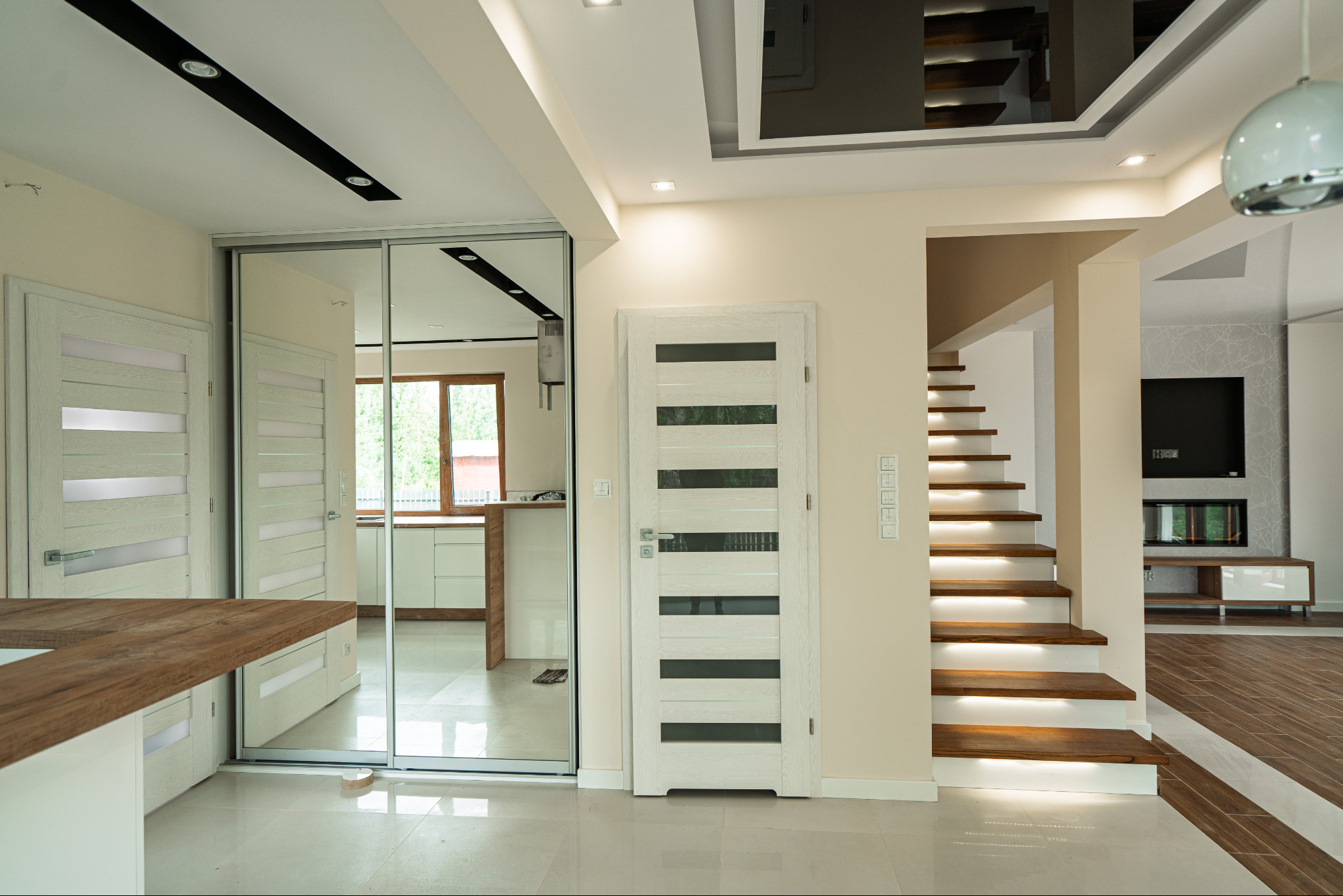Jasne wnętrze domu z drewnianymi schodami podświetlonymi LED, białymi drzwiami z przeszkleniami i lustrzaną szafą wnękową. Widoczna kuchnia w tle.