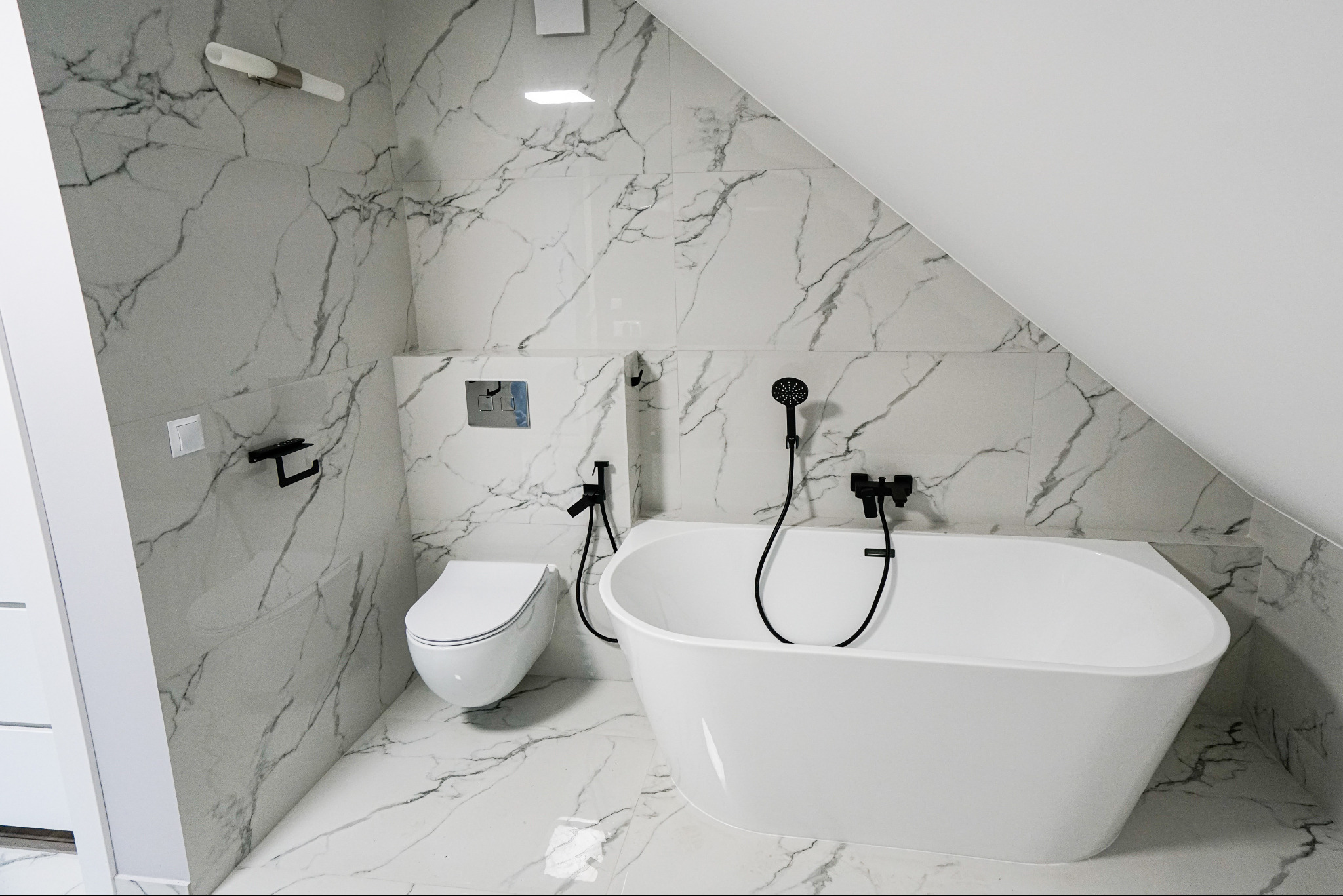 Biała, wolnostojąca wanna i toaleta w nowoczesnej łazience z marmurowymi płytkami. Czarna armatura i akcesoria. Poddasze ze skosem.