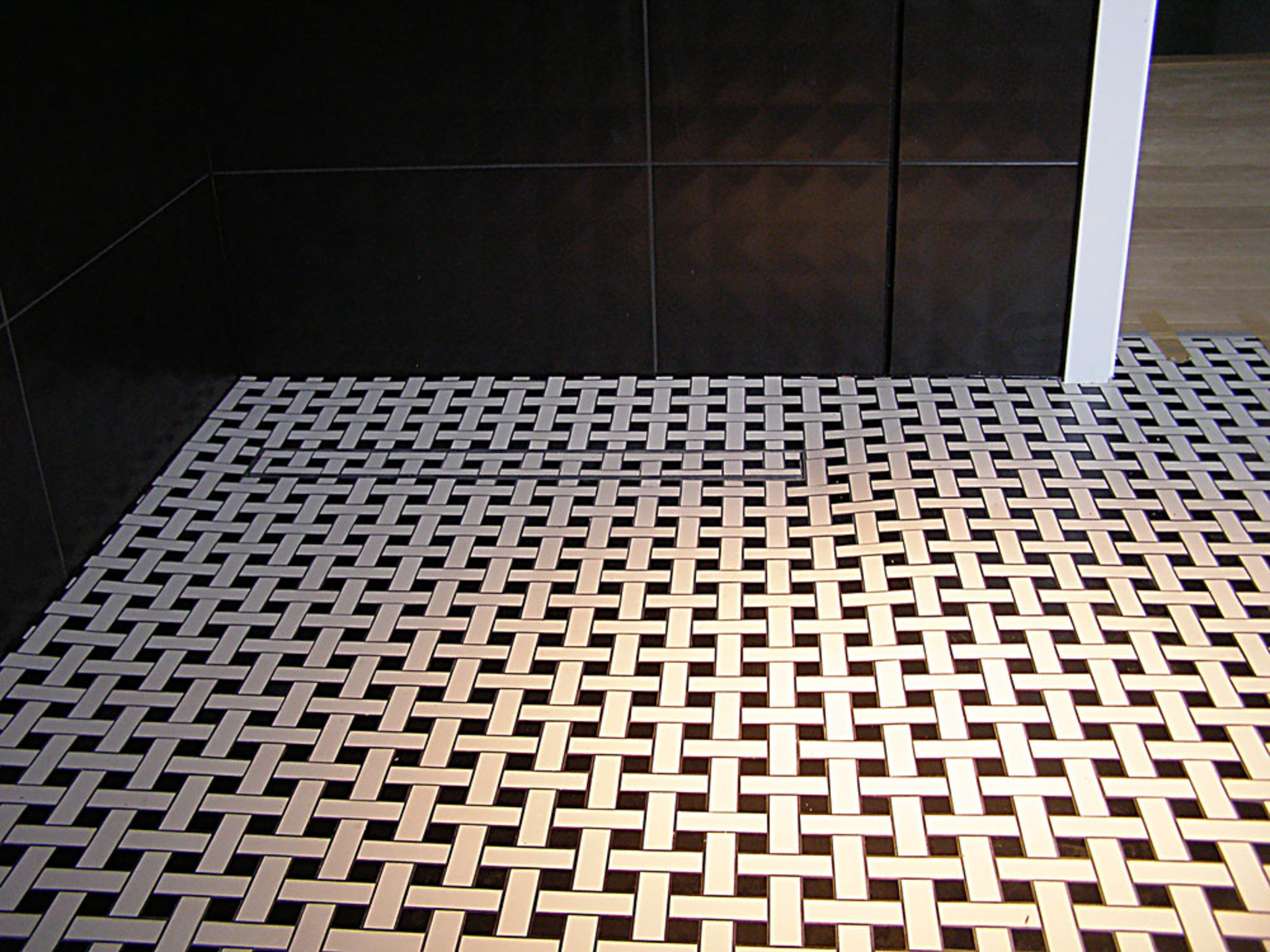 Ukończona podłoga w łazience z czarno-białą mozaiką ułożoną w wzór koszykowy. Na ścianie ciemne, duże płytki. Widoczny fragment białej listwy przypodłogowej.