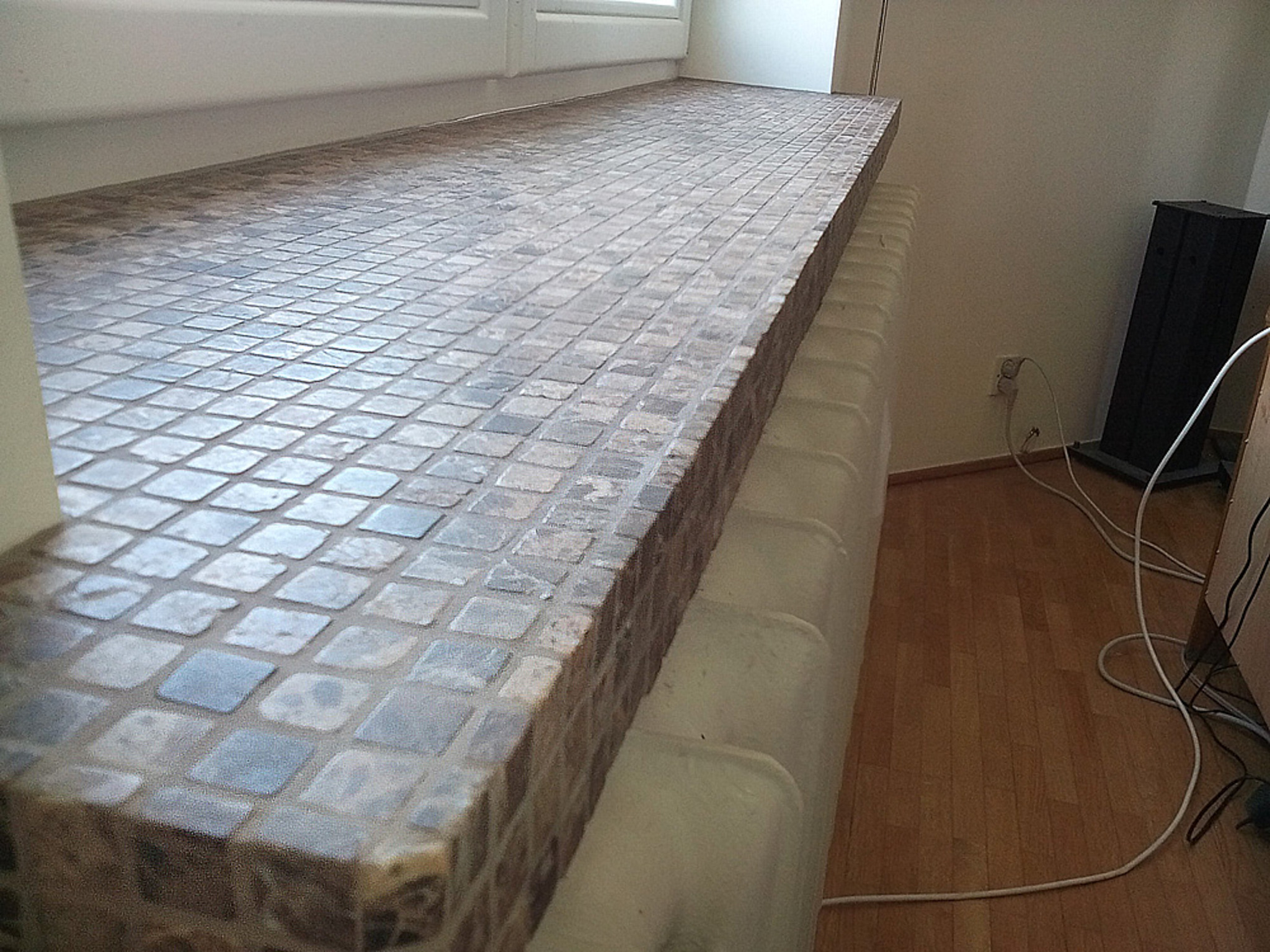 Mozaika z drobnych, kwadratowych płytek zdobi parapet okienny, przylegający do grzejnika. Widoczne drewniane panele podłogowe i fragment ściany z gniazdkiem.