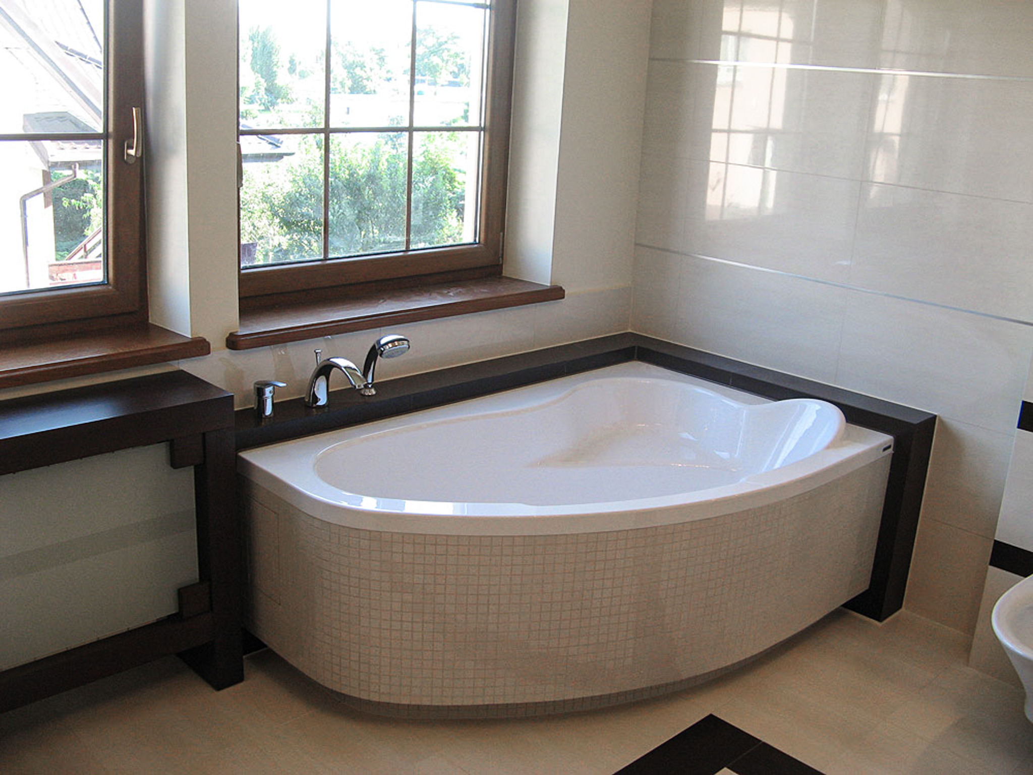 Nowoczesna łazienka z asymetryczną wanną obłożoną mozaiką, drewnianym oknem i beżowymi płytkami. Minimalistyczny design, jasne wnętrze z akcentami ciemnego drewna.