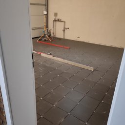 ZBIGNIEW USLUGI REMONTOWE - Układanie ciemnych płytek podłogowych w garażu, widoczne krzyżyki dystansowe, poziomica i lampa budowlana. Ujęcie zza uchylonych drzwi.