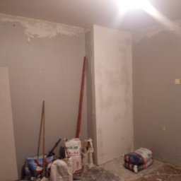 LUK-BUD - Pokój w trakcie prac wykończeniowych: częściowo wyszpachlowane ściany, worki z klejem i narzędzia malarskie. Widoczny fragment płyty karton-gips.