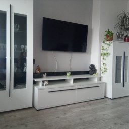 LUK-BUD - Nowoczesny salon z białymi meblami: szafka RTV, witryna z przeszkleniami, telewizor na ścianie, szara ściana i panele podłogowe. Minimalistyczny design z akcentami dekoracyjnymi.