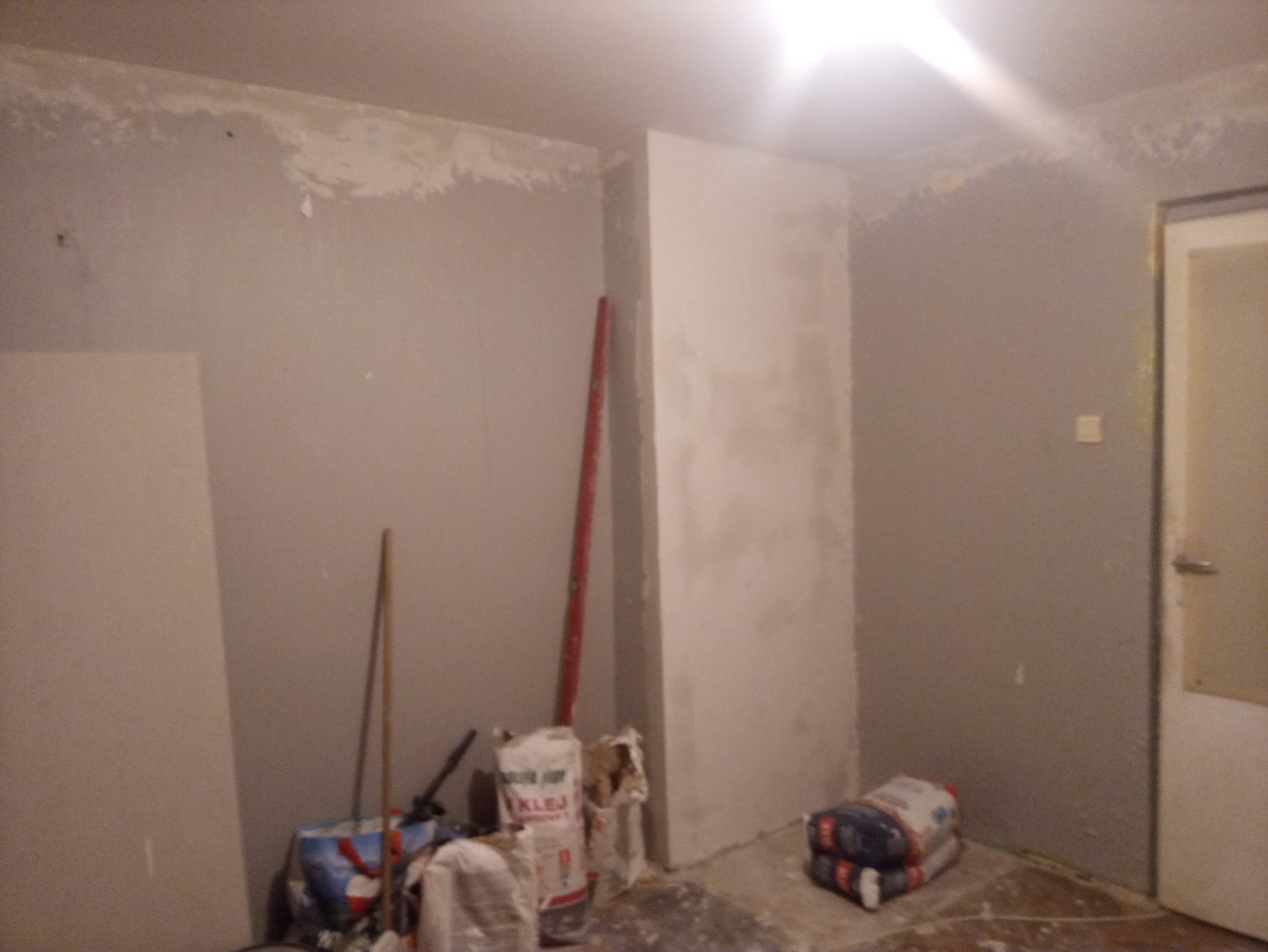 Pokój w trakcie prac wykończeniowych: częściowo wyszpachlowane ściany, worki z klejem i narzędzia malarskie. Widoczny fragment płyty karton-gips.