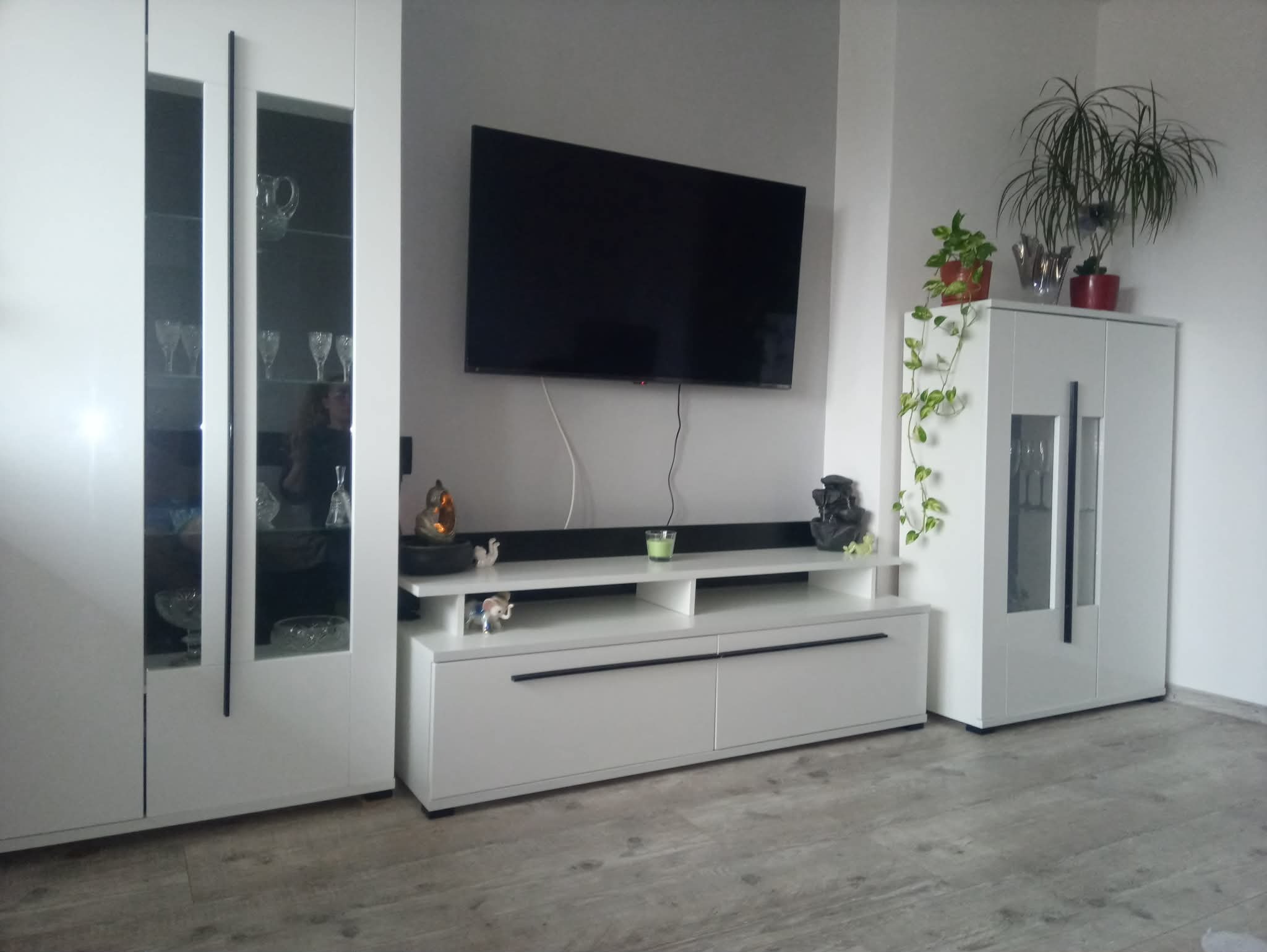 Nowoczesny salon z białymi meblami: szafka RTV, witryna z przeszkleniami, telewizor na ścianie, szara ściana i panele podłogowe. Minimalistyczny design z akcentami dekoracyjnymi.