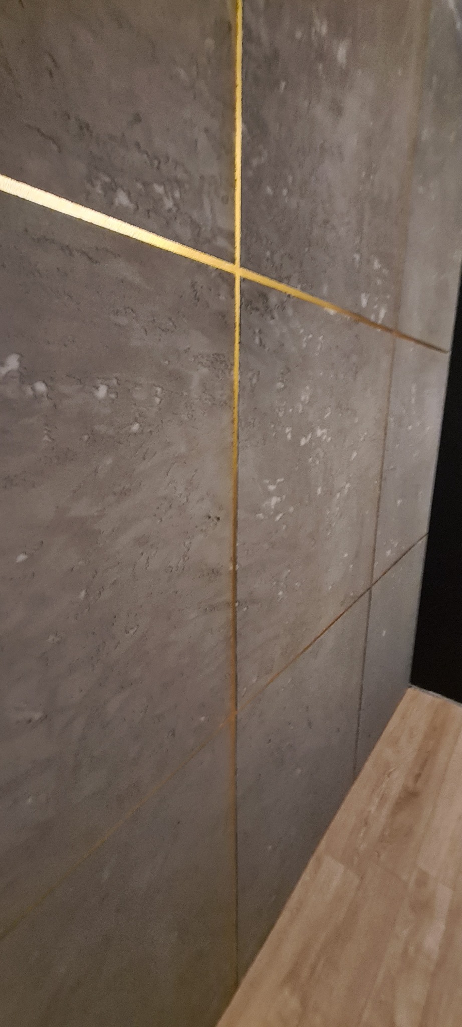 Szare płytki imitujące kamień z złotymi fugami krzyżowymi na ścianie, widoczne fragmenty podłogi z paneli. Ukończone wykończenie wnętrza.