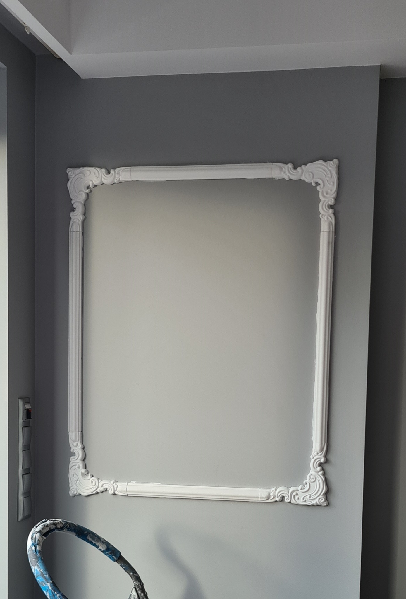 Szara ściana z dekoracyjną, białą ramą. W rogu widoczny fragment drabiny malarskiej. Detal wykończenia wnętrza w klasycznym stylu.