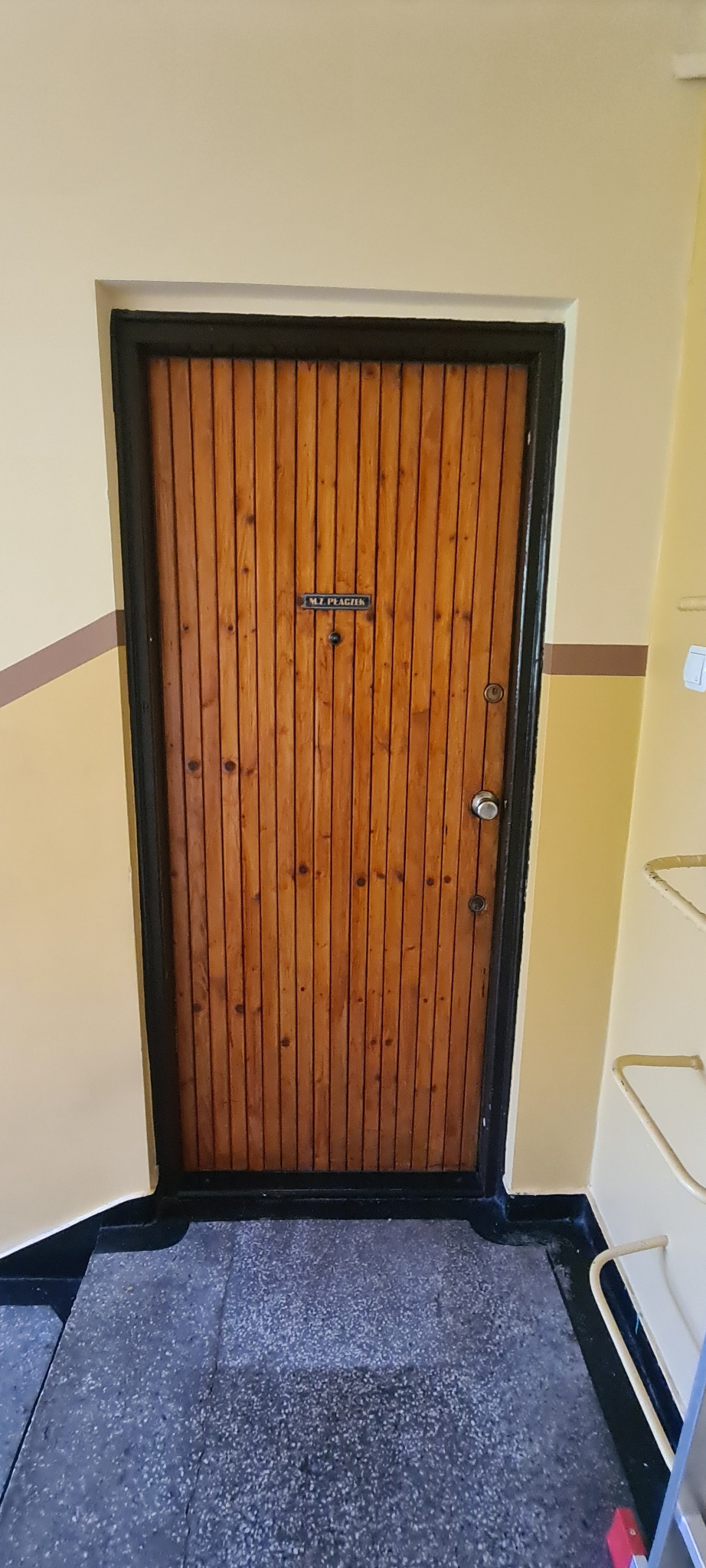 Drewniane drzwi wejściowe z czarną ramą w bloku mieszkalnym, widoczne schody i fragment ściany z brązowym paskiem. Na drzwiach tabliczka z nazwiskiem.