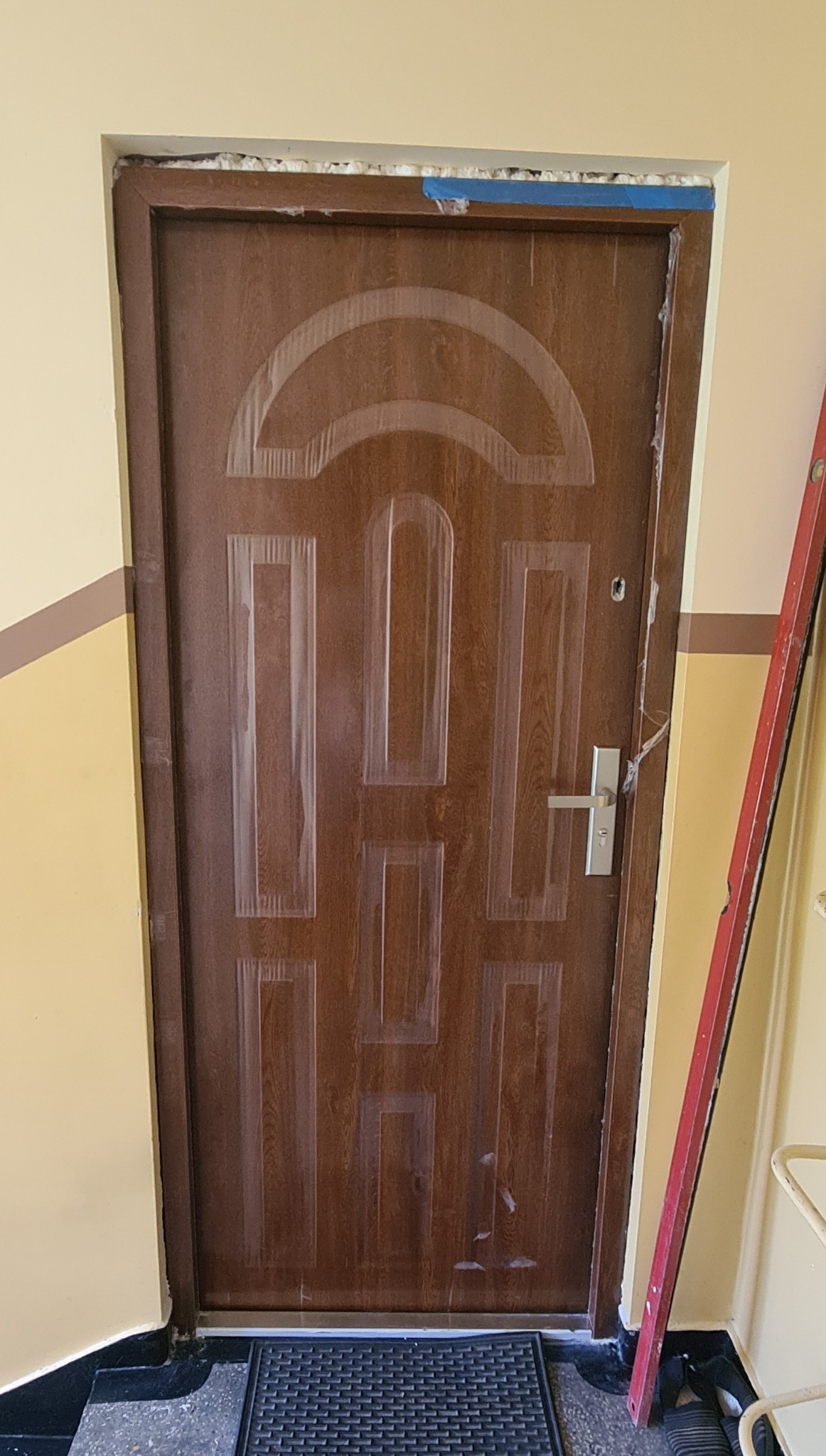 Brązowe drzwi z okleiną zabezpieczone folią malarską i taśmą, osadzone w ościeżnicy z widoczną pianką montażową. Poziomica oparta o ścianę.