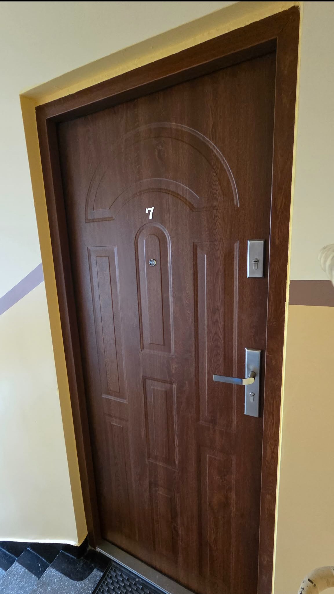 Ciemnobrązowe, drewnopodobne drzwi wejściowe z numerem '7' i srebrną klamką, zamontowane w żółtej futrynie na tle jasnej ściany klatki schodowej.