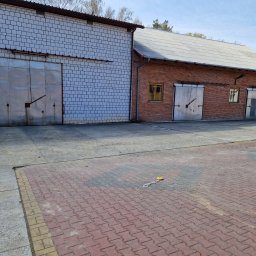 Wynajem chłodni - Dwa budynki magazynowe: biały z cegły silikatowej i czerwony z cegły, oba z metalowymi bramami. Na pierwszym budynku widoczne okno. Na ziemi leży żółty przedmiot.