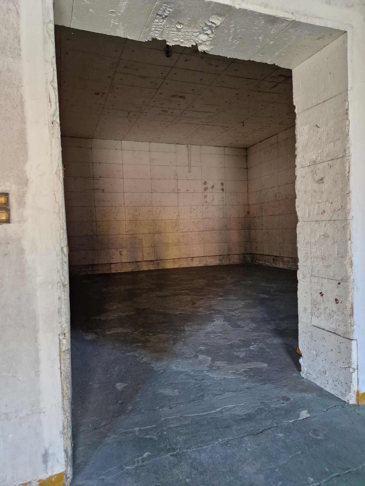 Wnętrze chłodni z betonowymi ścianami i podłogą, widoczne ślady wilgoci. Otwór wejściowy z surowym wykończeniem. Sufit z paneli. Widok z zewnątrz.