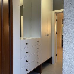 The Wood Life - Biała szafa wnękowa z szufladami i lustrem, brązowe uchwyty, orzechowe akcenty, dopasowana do przestrzeni, widok z boku, oświetlenie sufitowe, wnętrze domu.