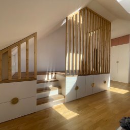 The Wood Life - Funkcjonalne łóżko na podeście ze schodami i szafkami w skosie poddasza, drewniana ażurowa ścianka działowa, jasna kolorystyka, okrągłe złote uchwyty.