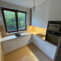 The Wood Life - Jasna kuchnia z białymi szafkami i drewnianym blatem, widok na zieleń za oknem. Uwagę przykuwa minimalistyczny design i ciepłe oświetlenie podszafkowe.