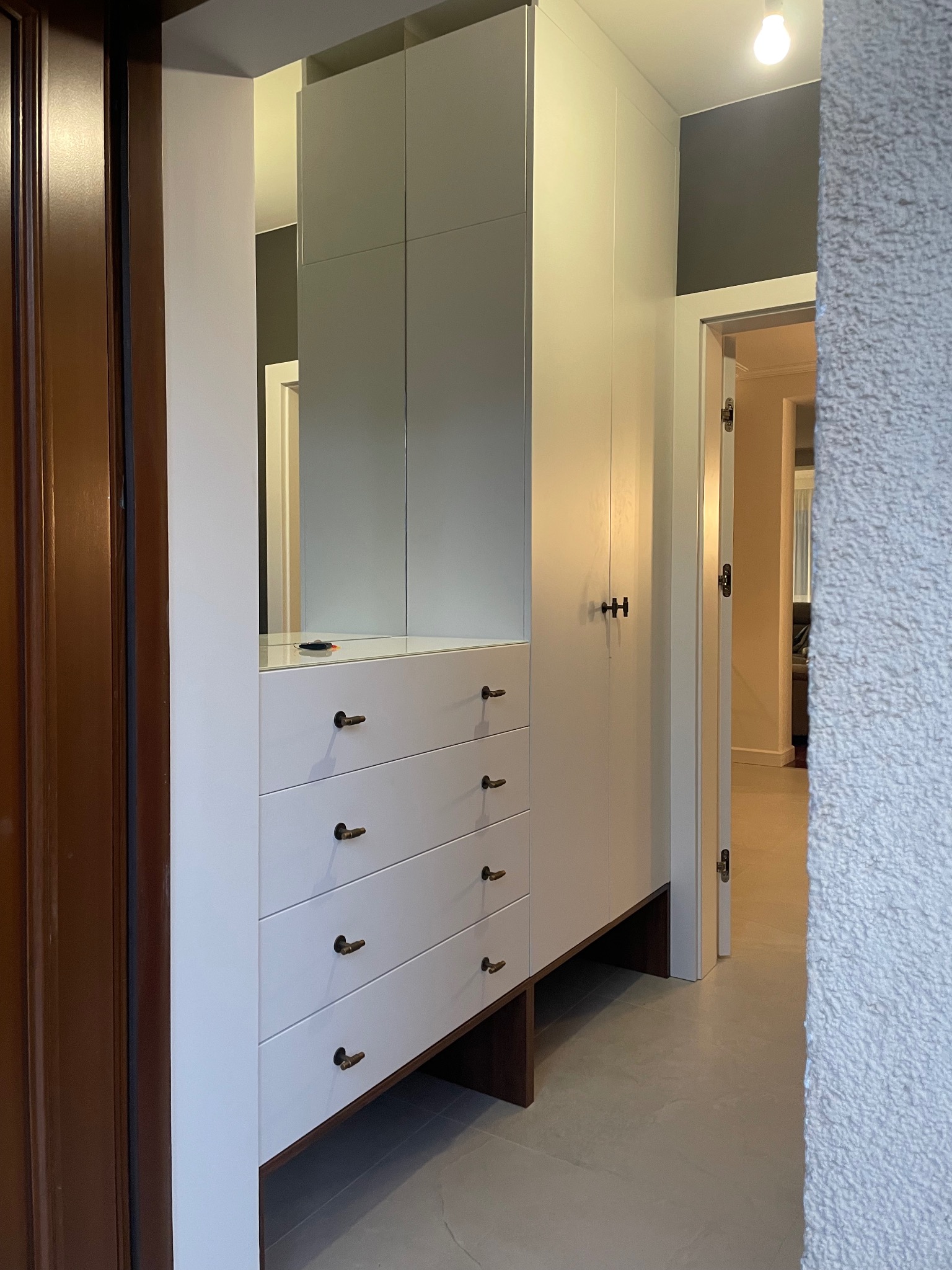 Biała szafa wnękowa z szufladami i lustrem, brązowe uchwyty, orzechowe akcenty, dopasowana do przestrzeni, widok z boku, oświetlenie sufitowe, wnętrze domu.