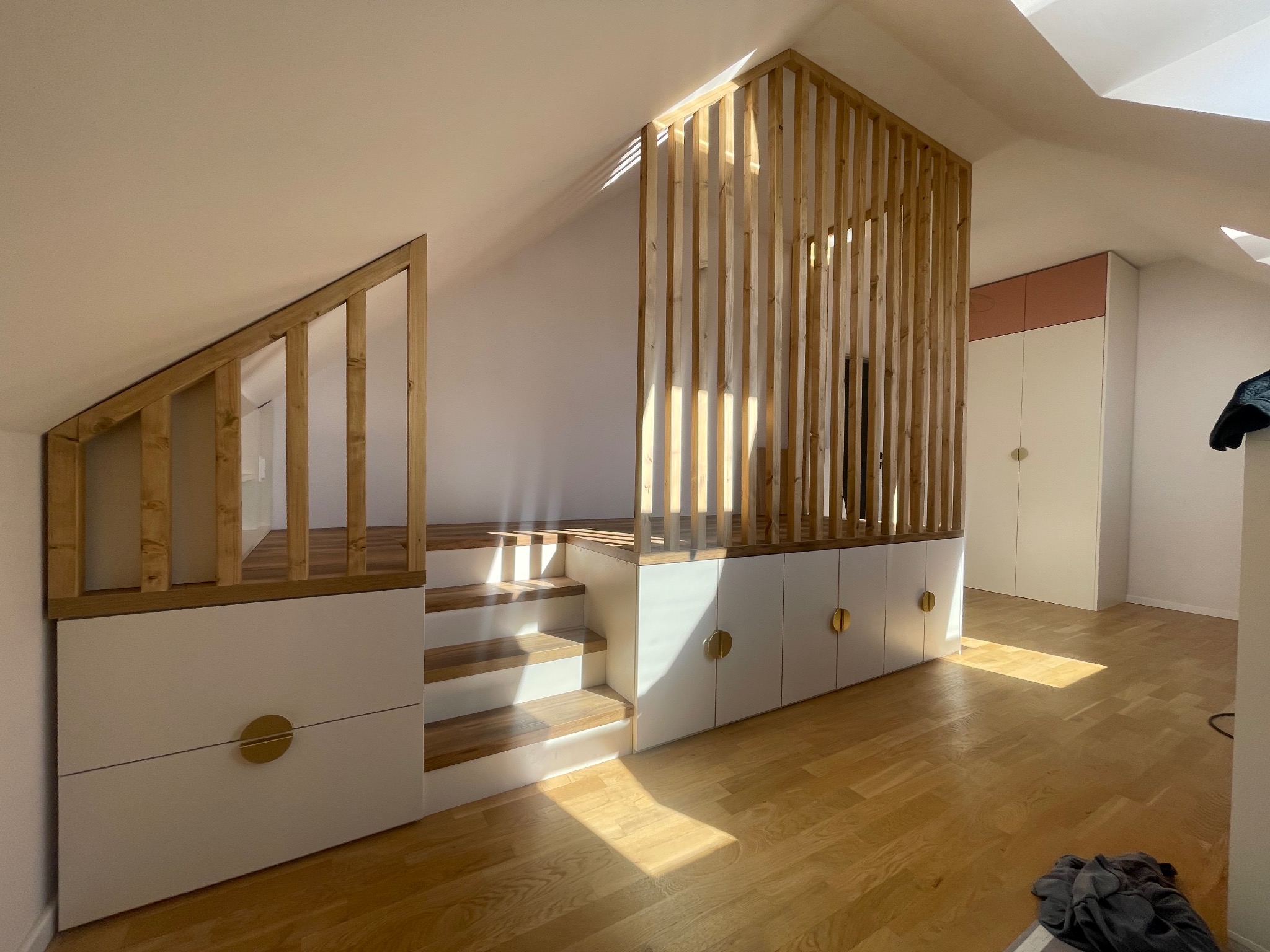 Funkcjonalne łóżko na podeście ze schodami i szafkami w skosie poddasza, drewniana ażurowa ścianka działowa, jasna kolorystyka, okrągłe złote uchwyty.