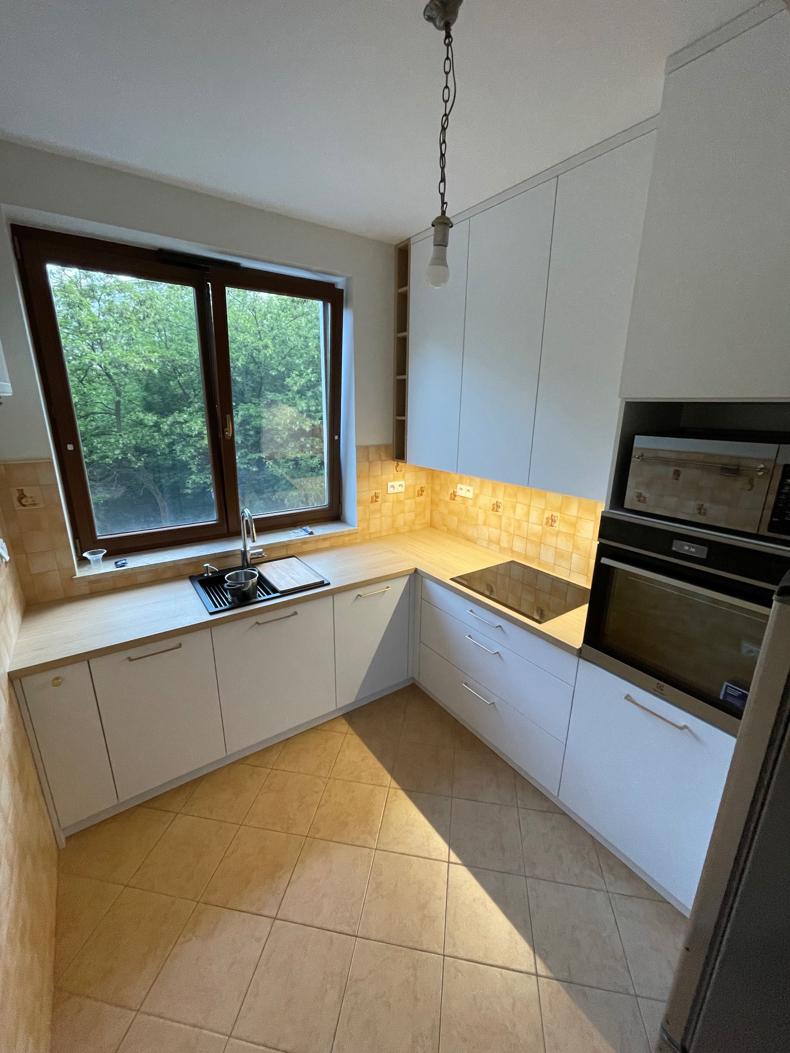 Jasna kuchnia z białymi szafkami i drewnianym blatem, widok na zieleń za oknem. Uwagę przykuwa minimalistyczny design i ciepłe oświetlenie podszafkowe.