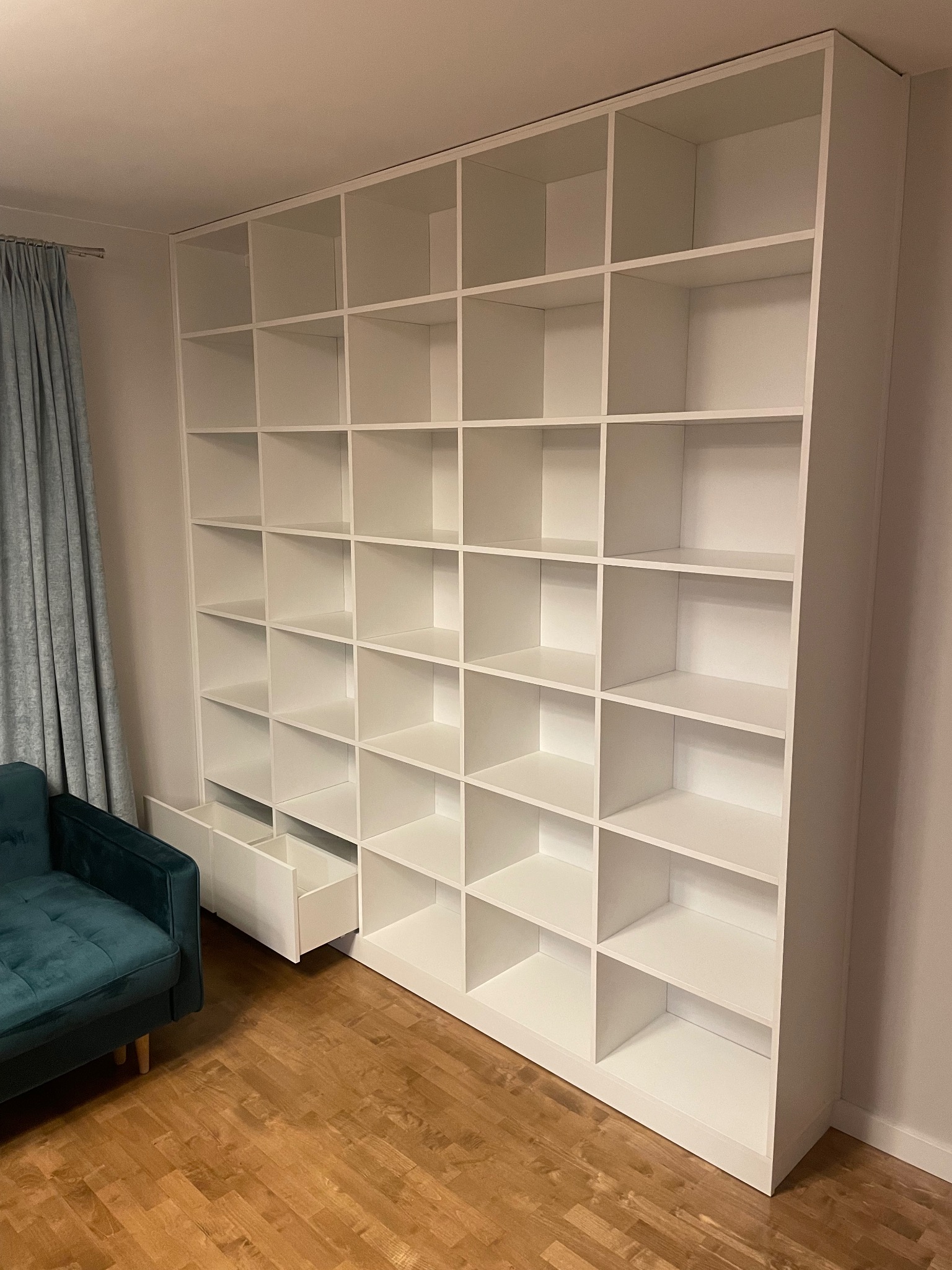 Biała, duża szafa wnękowa z otwartymi półkami i wysuwanymi szufladami, dopasowana do sufitu, widoczna z boku, obok stoi zielony fotel.