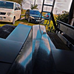 Pi3GuS-DrEwNoTyP - Błyszczące, czarne arkusze blachy na pierwszym planie, w tle drabina, samochody i błękitne niebo z chmurami. Nietypowa perspektywa.