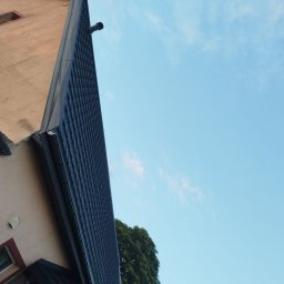 Pi3GuS-DrEwNoTyP - Ciemna dachówka ceramiczna na skośnym dachu domu jednorodzinnego, widok z dołu na tle błękitnego nieba z chmurami i zieleni drzewa. Rynna w kolorze dachu.