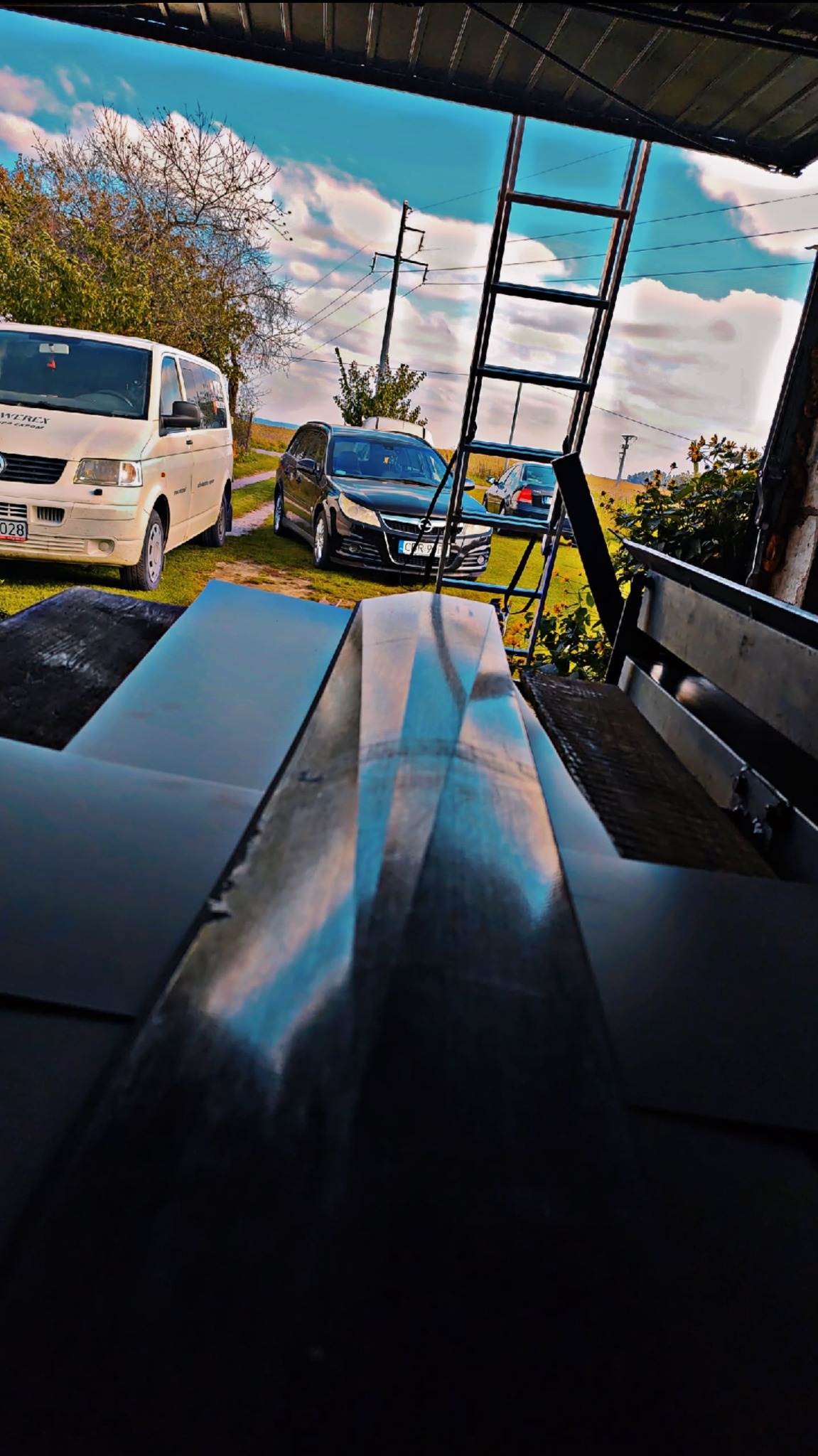 Błyszczące, czarne arkusze blachy na pierwszym planie, w tle drabina, samochody i błękitne niebo z chmurami. Nietypowa perspektywa.