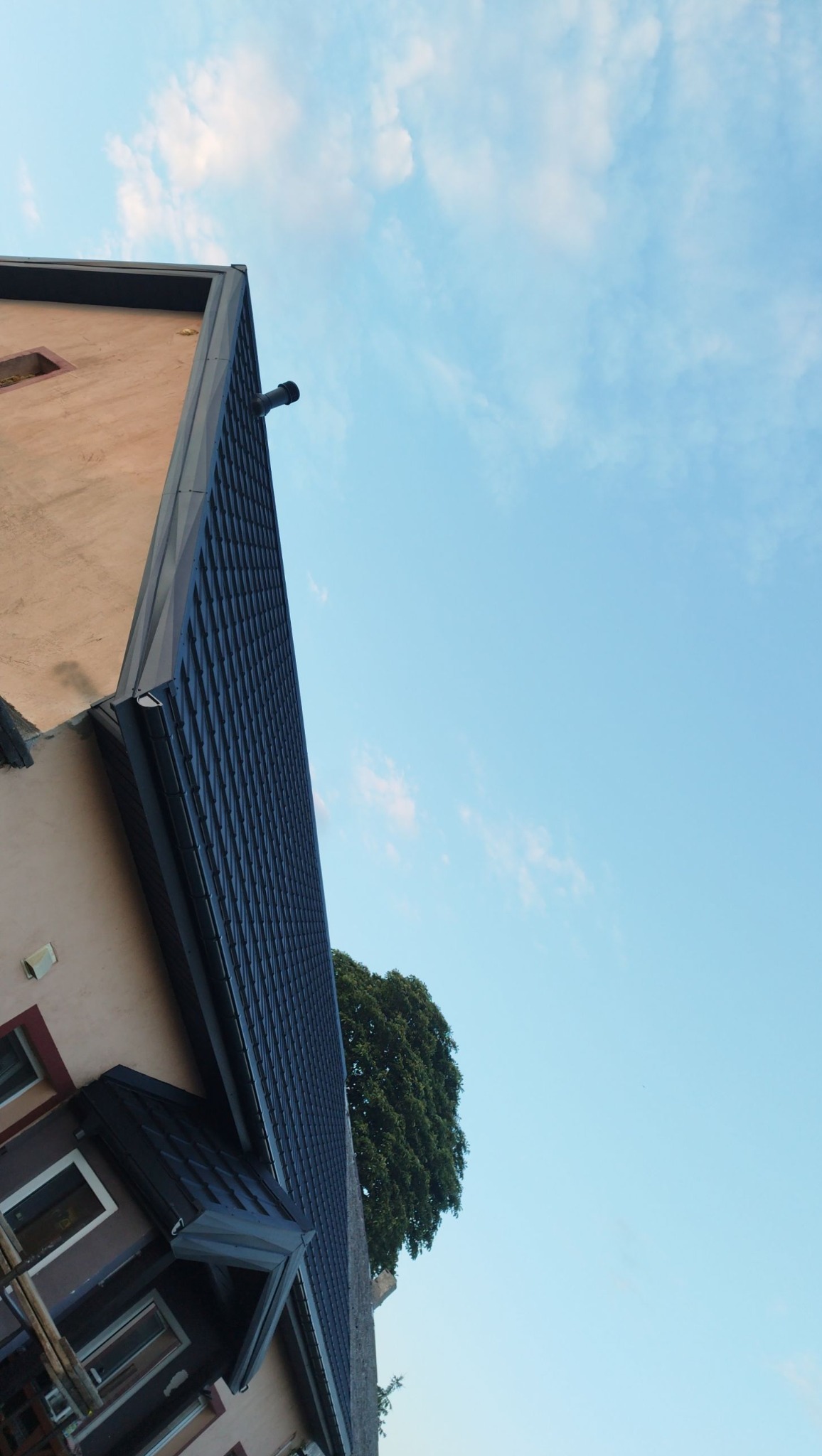 Ciemna dachówka ceramiczna na skośnym dachu domu jednorodzinnego, widok z dołu na tle błękitnego nieba z chmurami i zieleni drzewa. Rynna w kolorze dachu.