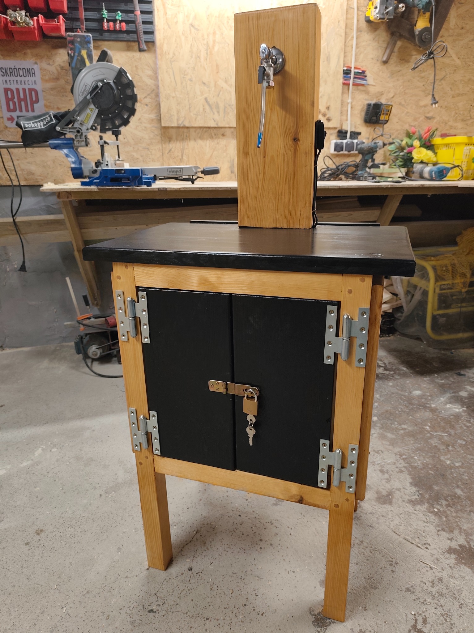 Drewniana szafka warsztatowa z czarnymi drzwiczkami, zabezpieczona zamkiem na klucz. Na blacie zamontowany drewniany element z kranem i wężykiem. W tle narzędzia.