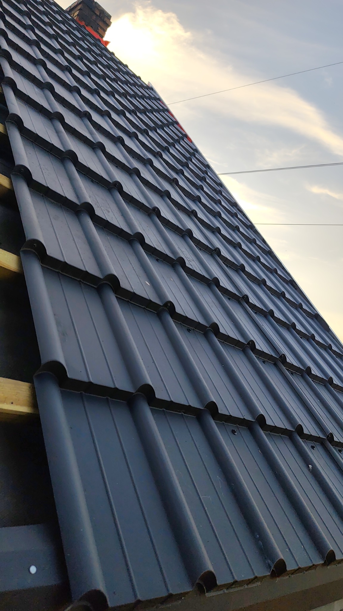 Nowe, grafitowe pokrycie dachowe z blachy trapezowej, widoczne drewniane łaty, fragment komina w tle, pochmurne niebo. Ujęcie z dołu pod kątem.