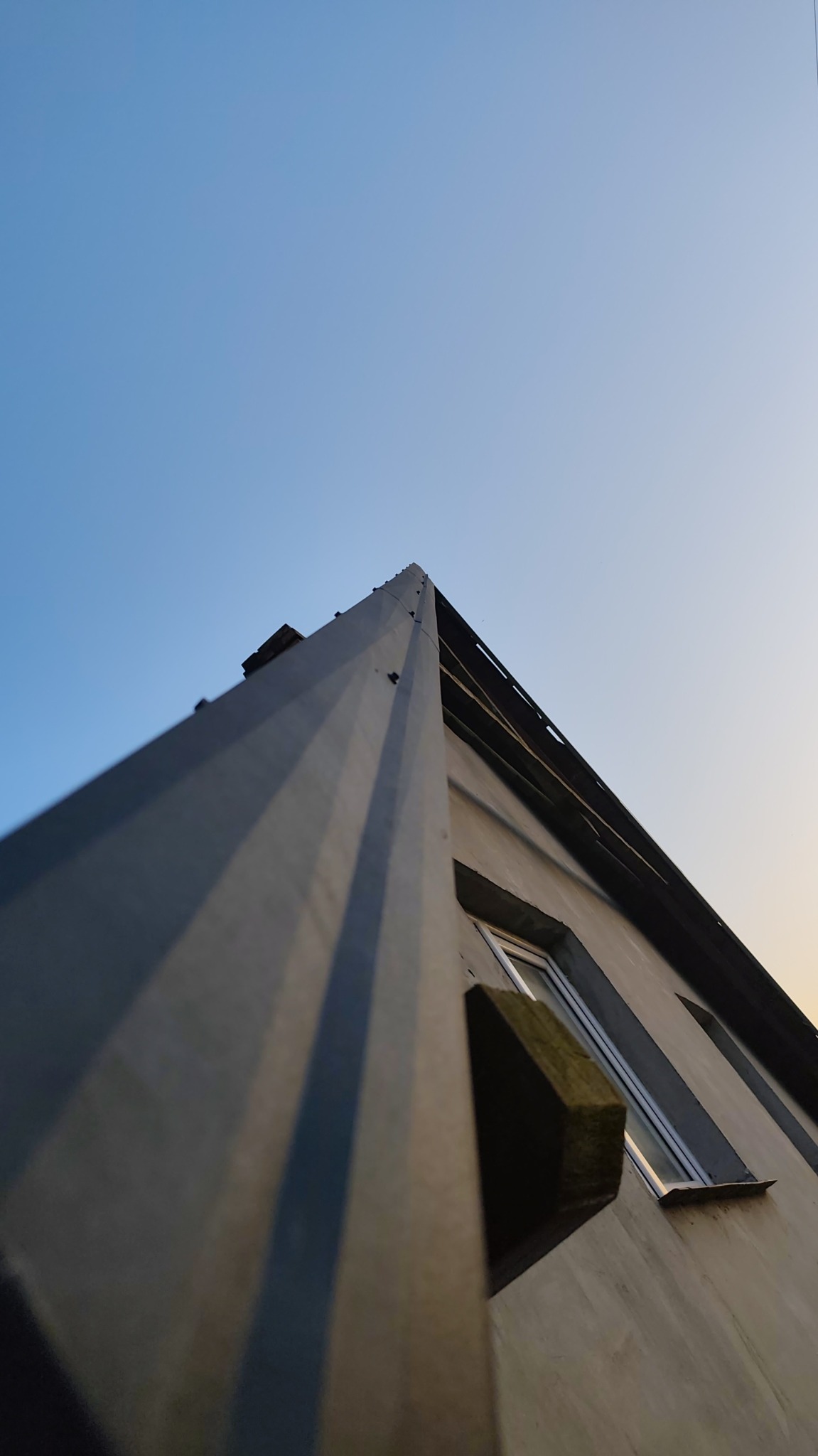 Nietypowe ujęcie pionowe na szczyt budynku z szarym dachem i fragmentem ściany z oknem na tle błękitnego nieba o zmierzchu. Perspektywa z dołu.