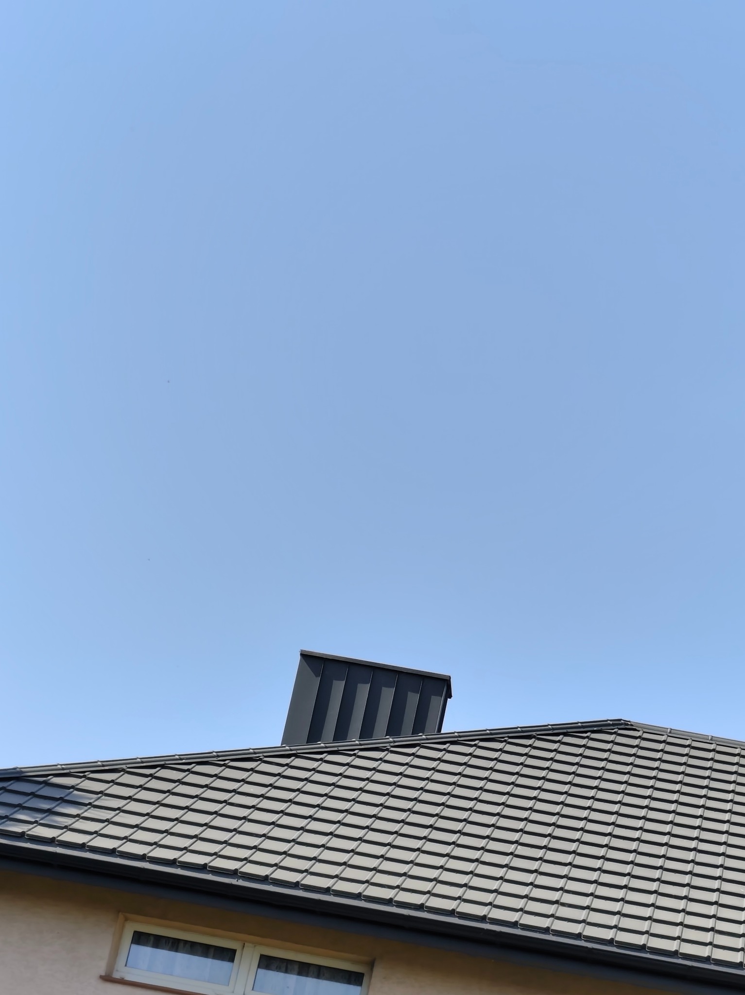 Szary dach z dachówki ceramicznej i kominem wentylacyjnym na tle błękitnego nieba. Widoczny fragment elewacji budynku z oknem.
