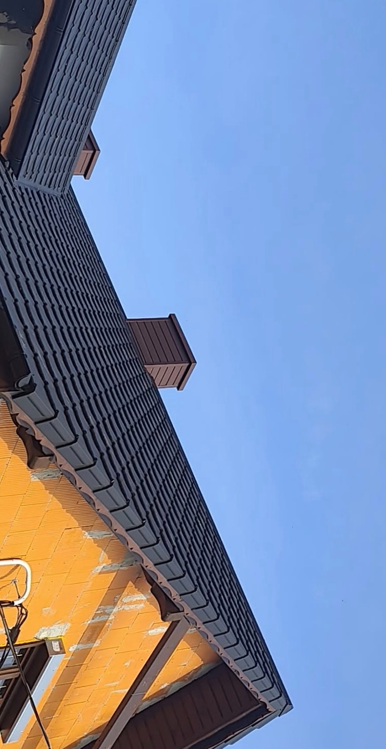 Ciemna dachówka ceramiczna na budynku w trakcie budowy, widoczny komin i fragment ściany z pustaków. Ujęcie pod kątem, błękitne niebo w tle.