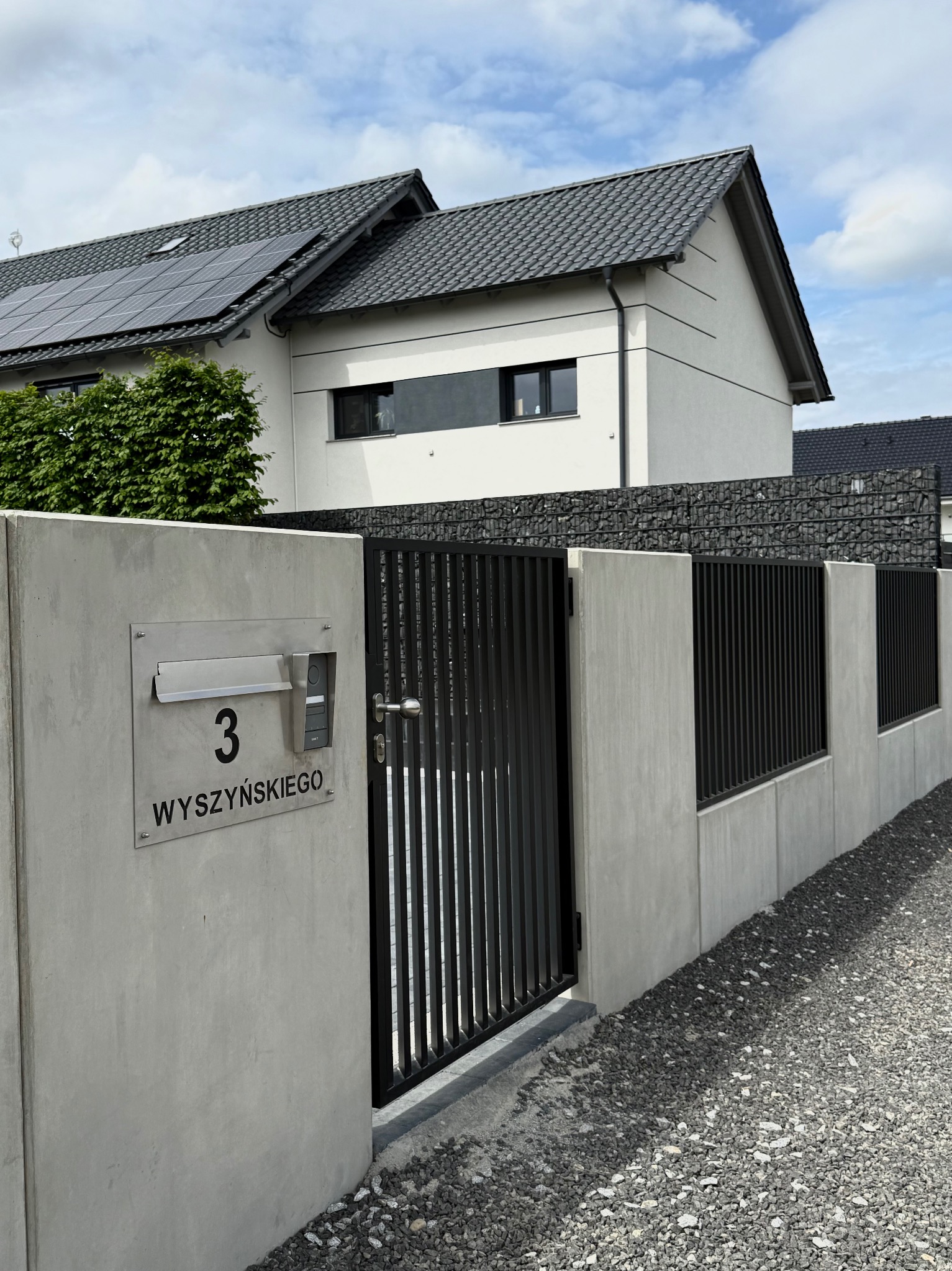 Nowoczesne ogrodzenie z metalową bramą i betonowymi słupkami, uzupełnione gabionami. Widoczny numer posesji i dom w tle.