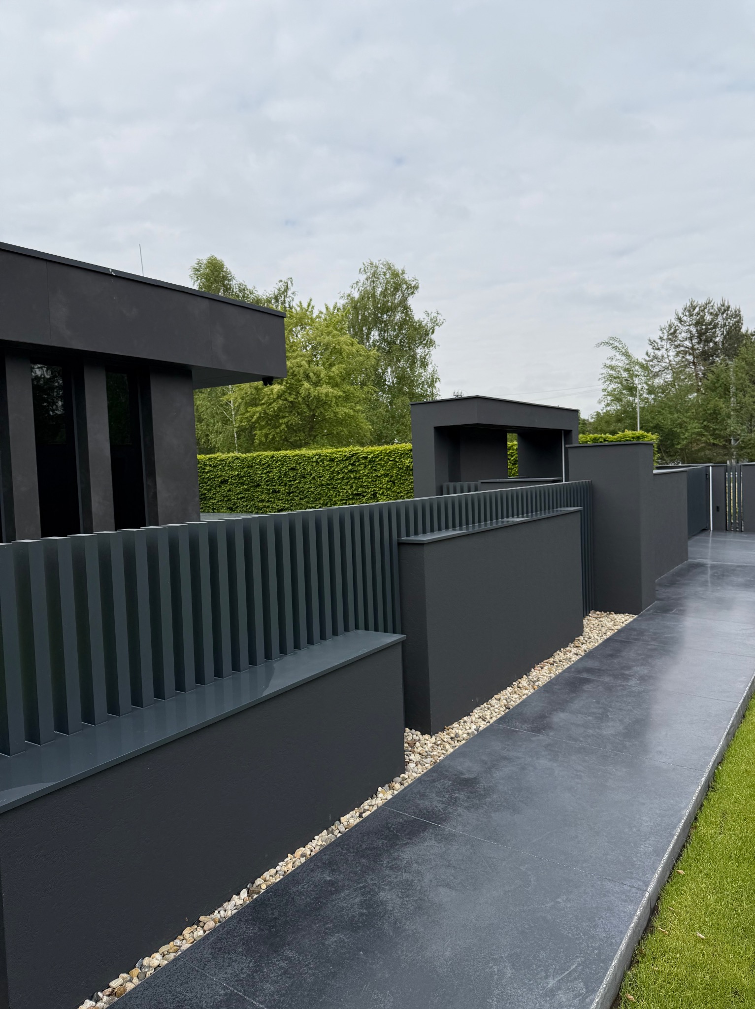 Nowoczesne ogrodzenie metalowe w kolorze antracytowym z pionowymi lamelami i elementami murowanymi, oddzielone od chodnika drobnymi kamieniami.