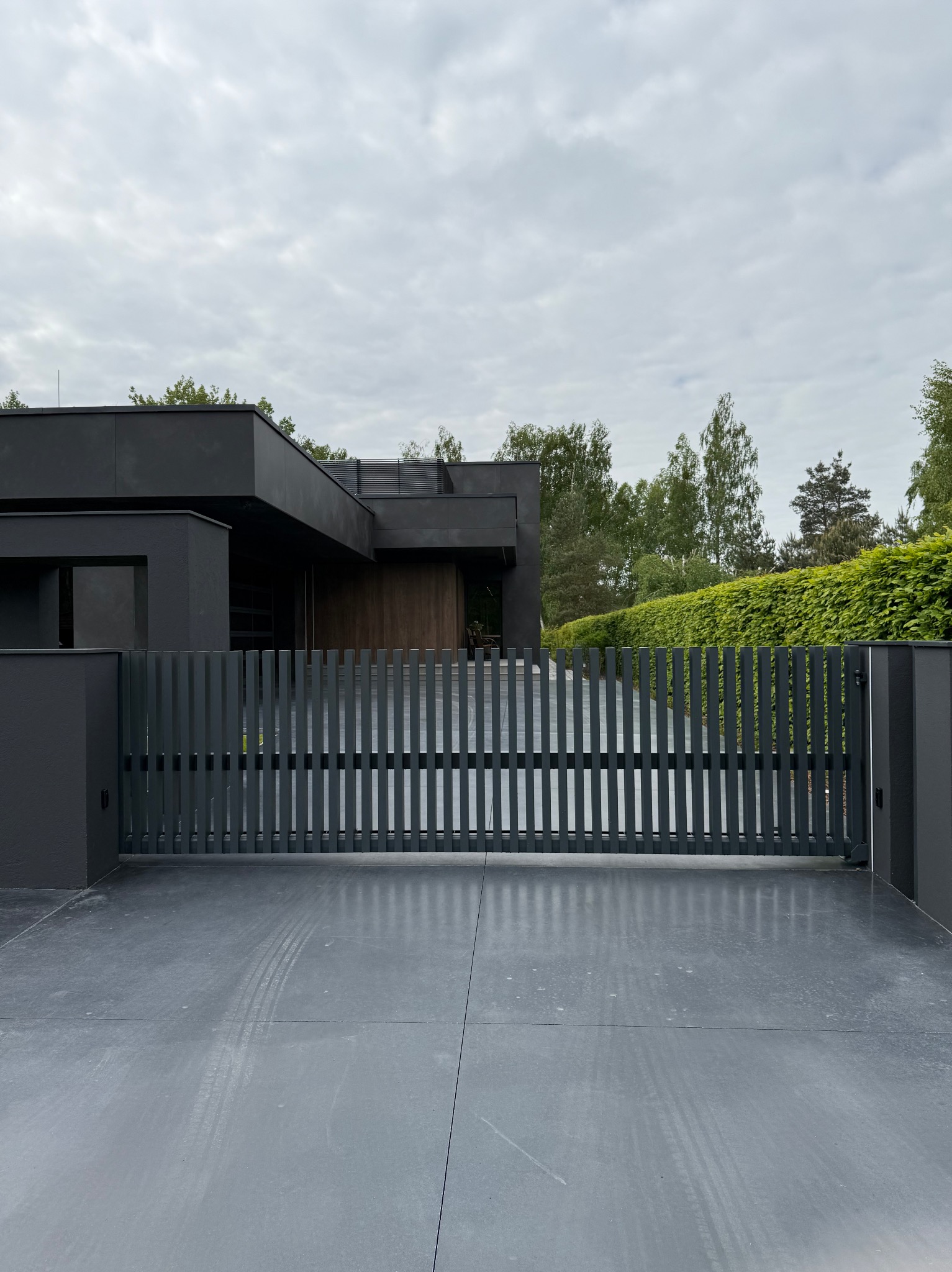 Nowoczesna, przesuwna brama w kolorze grafitowym z pionowymi szczeblami, zamontowana przed minimalistycznym domem z płaskim dachem i zadbanym żywopłotem w tle.