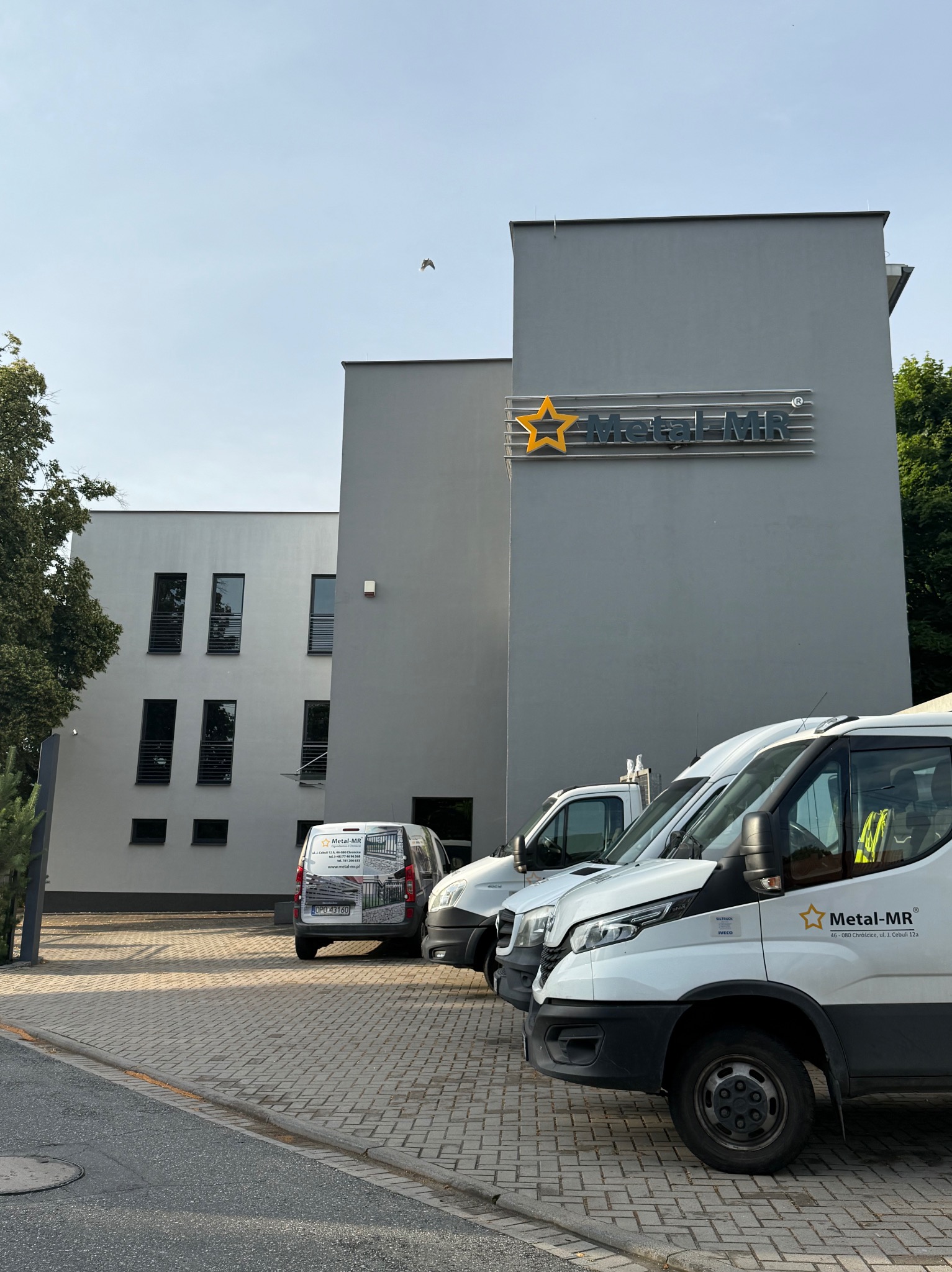 Szary budynek firmy Metal-MR z logo i zaparkowanymi samochodami dostawczymi z logo firmy. Widok z poziomu ulicy, w tle drzewa i błękitne niebo.