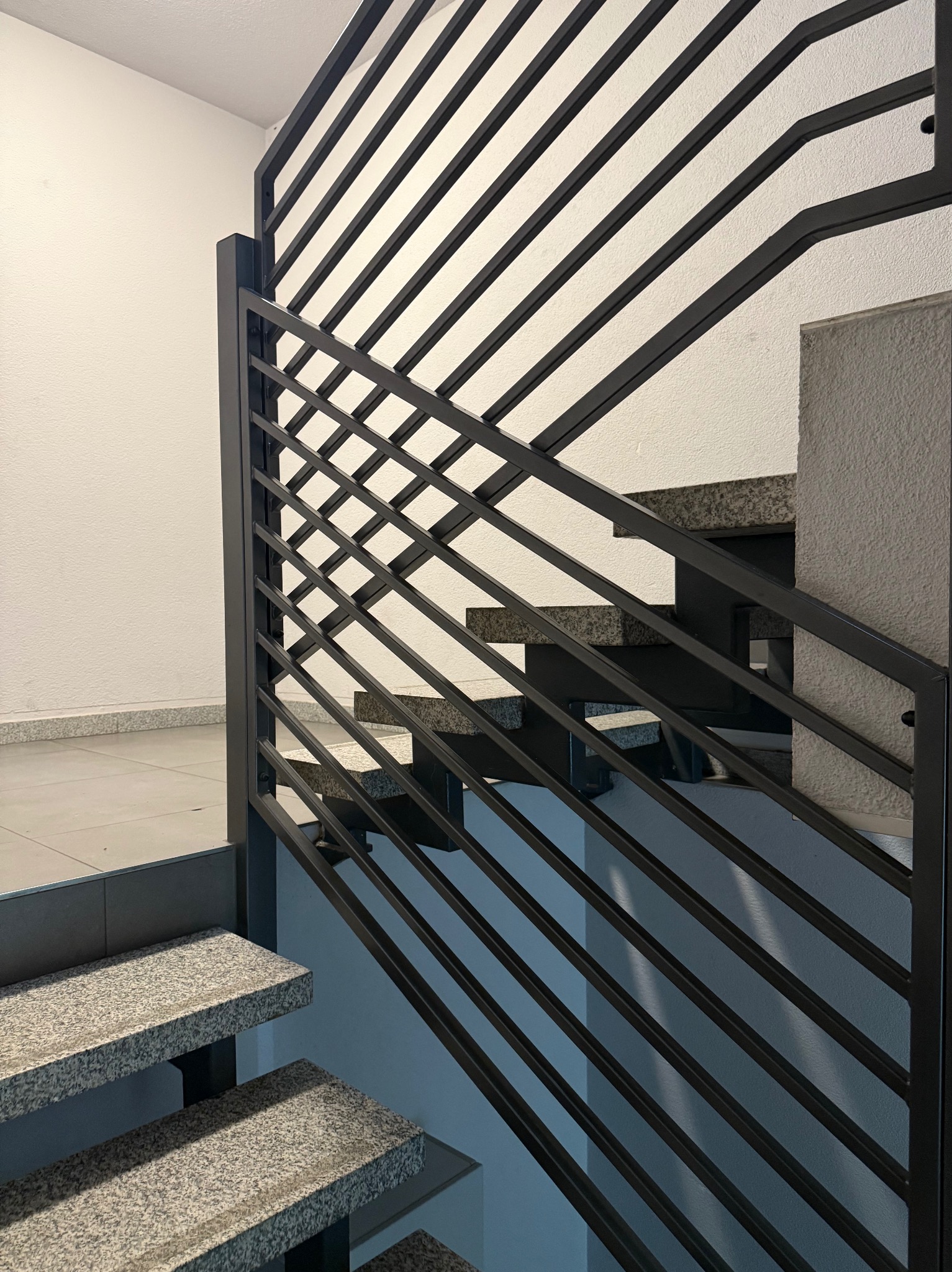 Nowoczesne schody z szarego granitu z czarną, metalową balustradą o geometrycznym wzorze. Minimalistyczny design, jasne wnętrze, widok z dołu na górę.