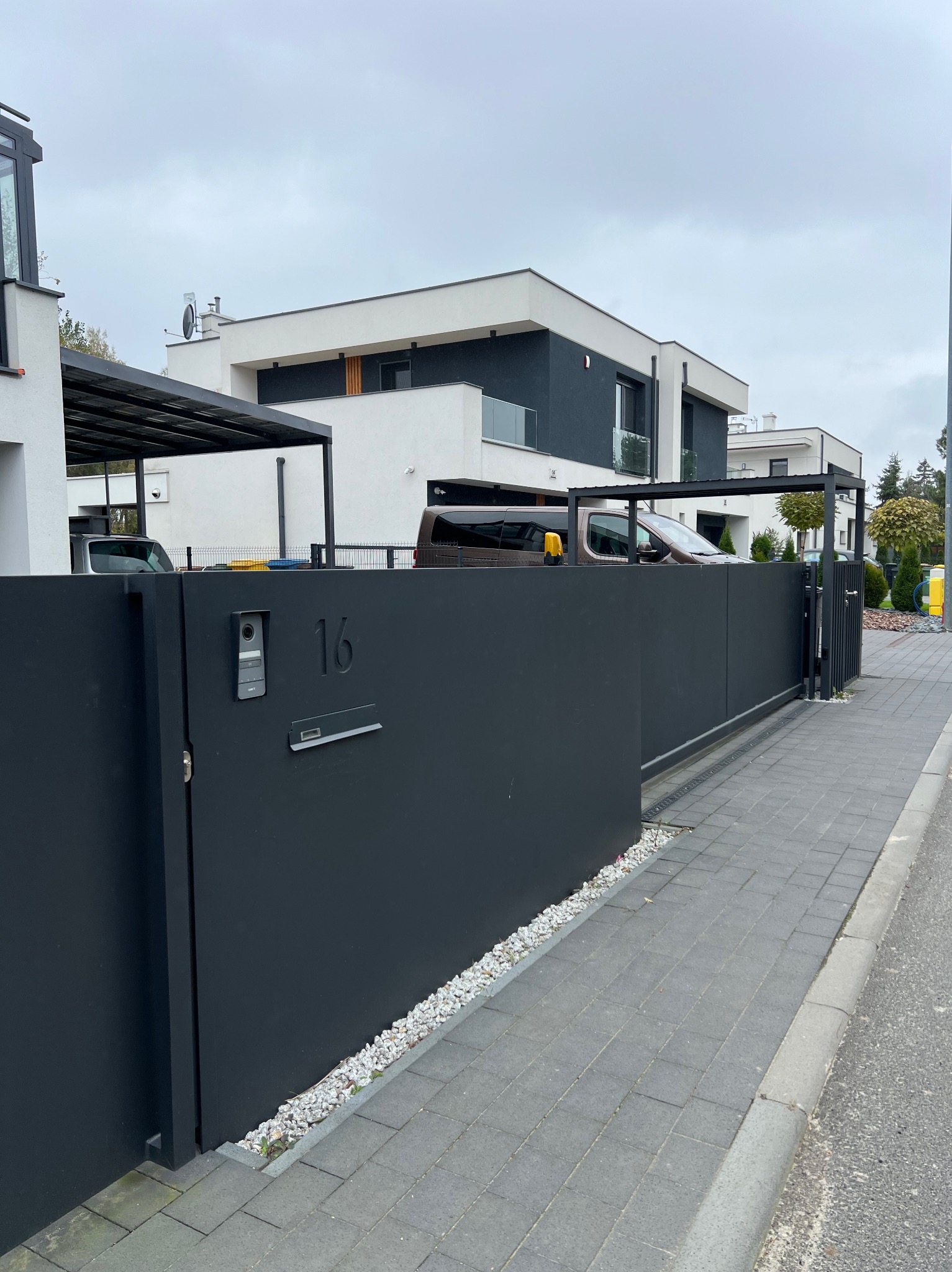 Nowoczesna, przesuwna brama w kolorze antracytu z numerem domu i domofonem, na tle minimalistycznego budynku mieszkalnego i szarej kostki brukowej.