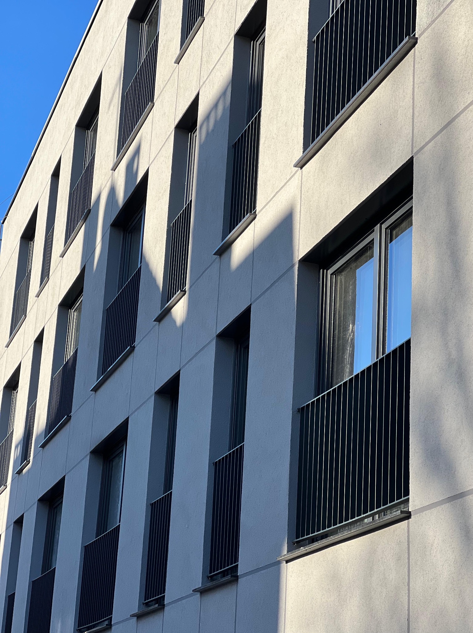 Fasada budynku z balkonami zabezpieczonymi metalowymi, pionowymi balustradami. Widok z dołu, podkreślony kontrastem światła i cienia.