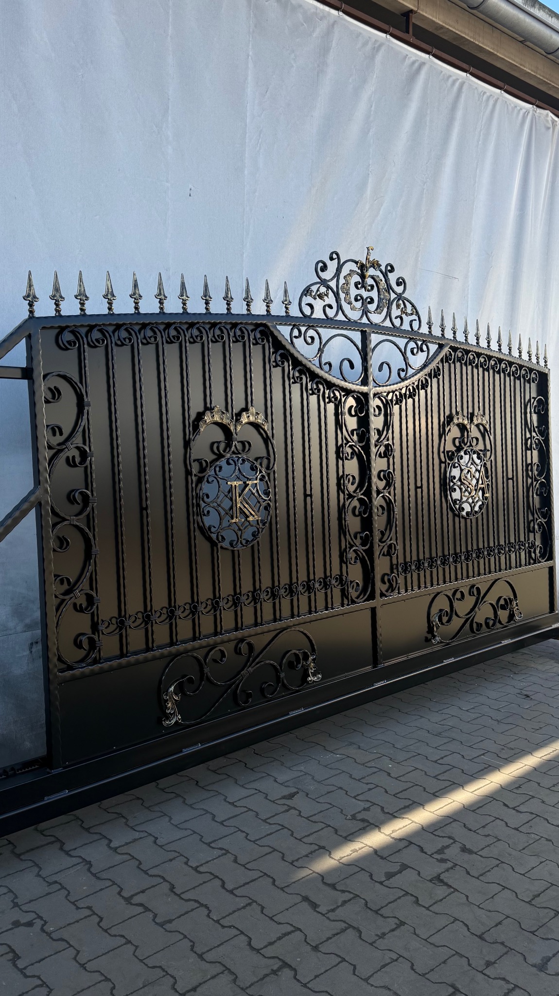 Czarna, metalowa brama przesuwna z dekoracyjnymi elementami i monogramami, ustawiona na kostce brukowej przed białym płótnem. Kute zdobienia i szpiczaste zakończenia.
