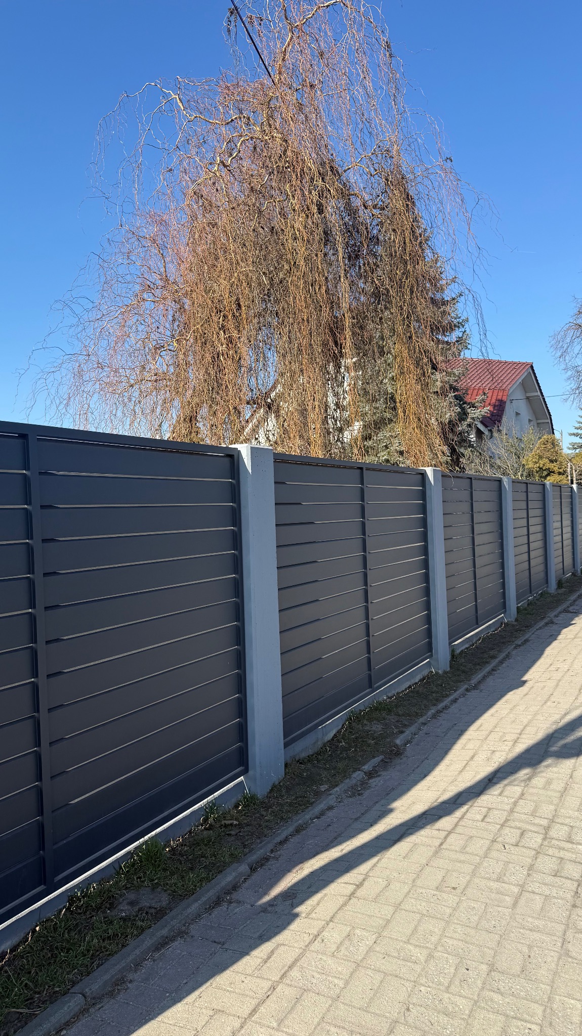 Nowoczesne ogrodzenie posesji z poziomych, grafitowych paneli, osadzone na betonowych słupkach. W tle drzewo i dom z czerwonym dachem w słoneczny dzień.