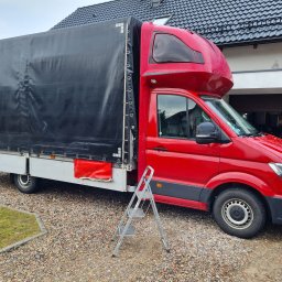 Rafał Rebicki Firma Handlowo Szkoleniowa err - Czerwony bus z czarną plandeką zaparkowany na żwirowym podjeździe, z rozłożoną drabiną obok. Widoczny fragment domu z garażem w tle.