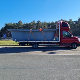 Rafał Rebicki Firma Handlowo Szkoleniowa err - Czerwony bus z kabiną sypialną przewozi szary, duży zbiornik zabezpieczony pasami transportowymi. Widok z boku, w tle drzewa i błękitne niebo.